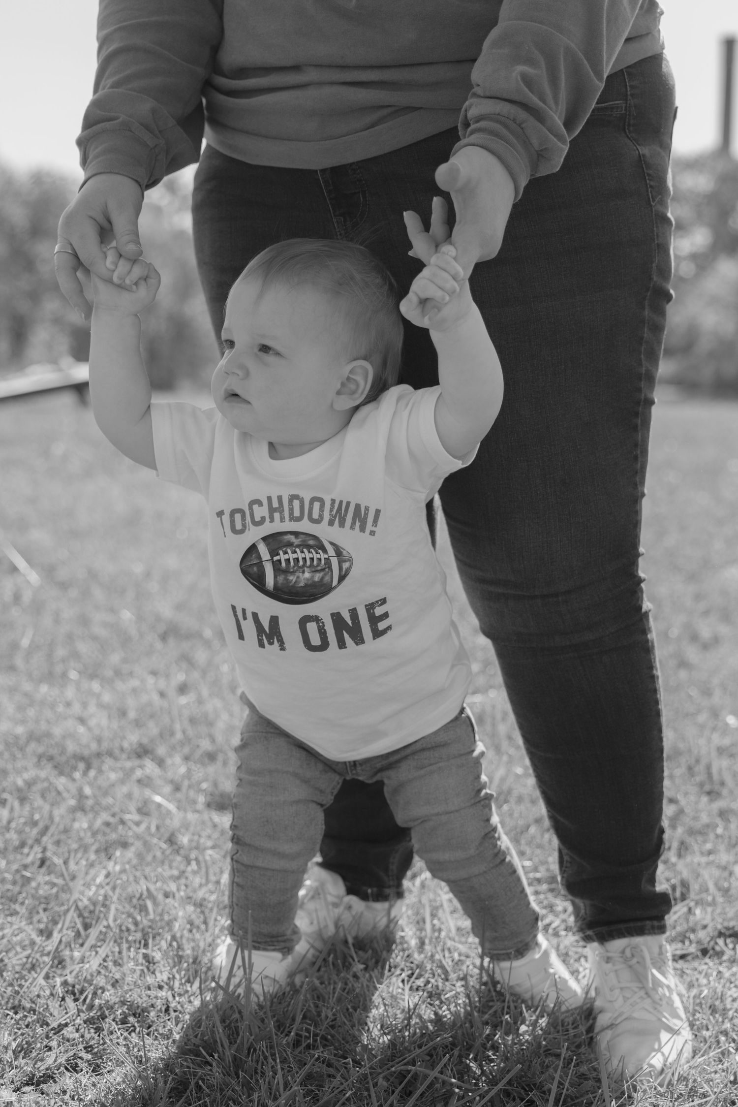 Black and white photo series of a baby learning to walk while wearing a football-themed onesie in a grassy field.