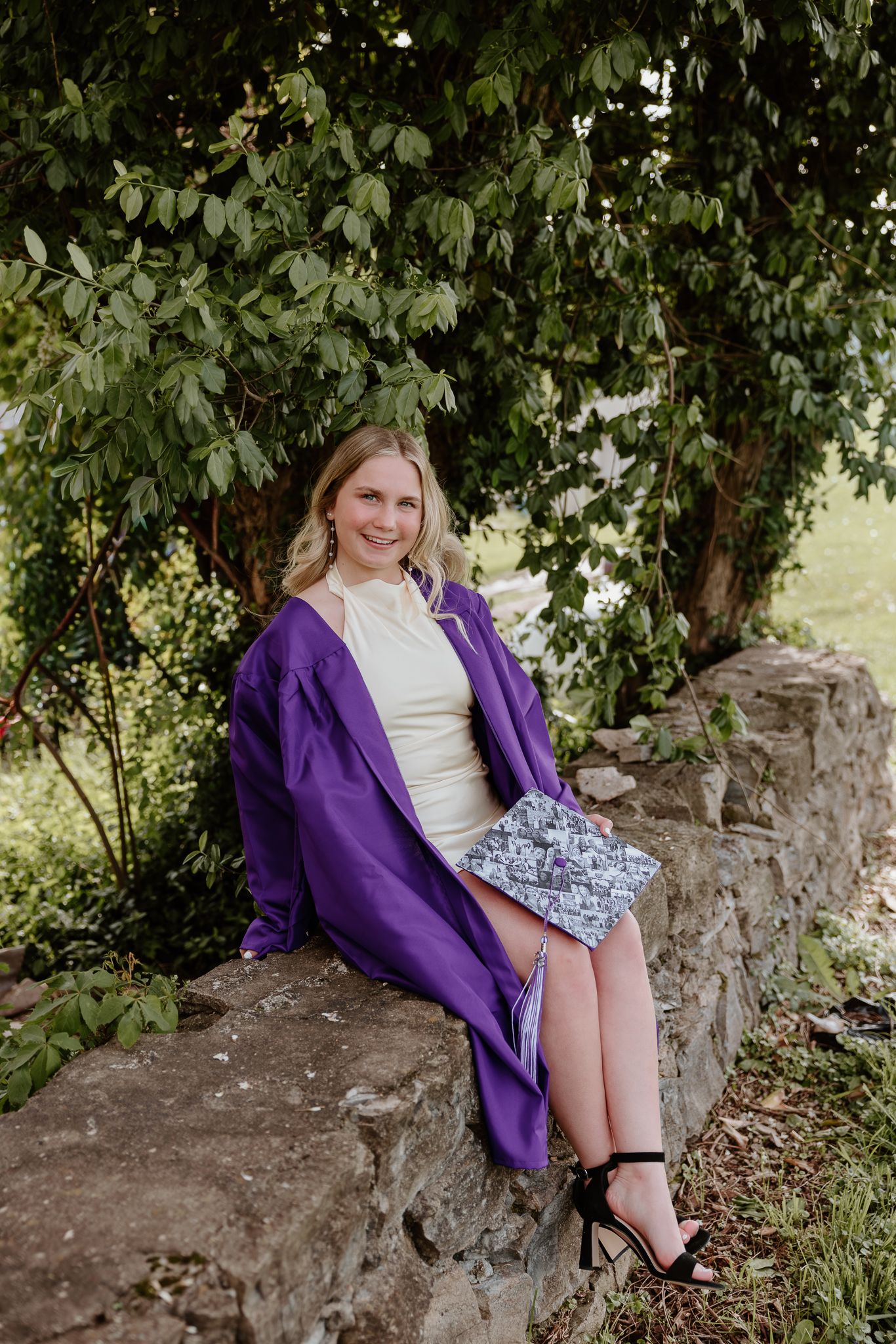 Young woman in purple graduation gown and cream dress sits on stone wall outdoors, smiling, holding decorative clutch.