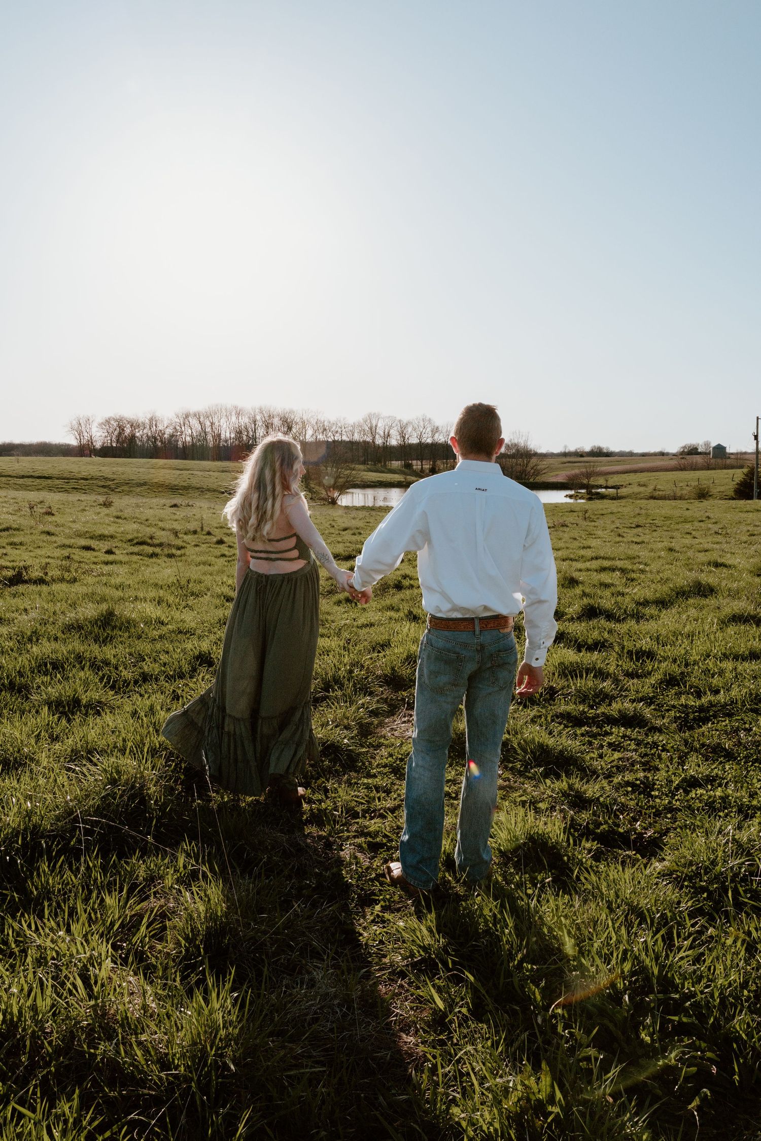 Two people walk hand in hand through a sunlit green meadow at sunset with trees on the horizon.