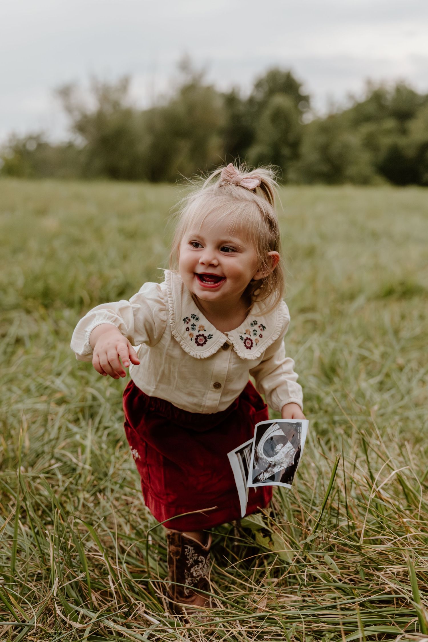 A small child in a white collared top and red pants plays joyfully in tall grass while holding photographs.