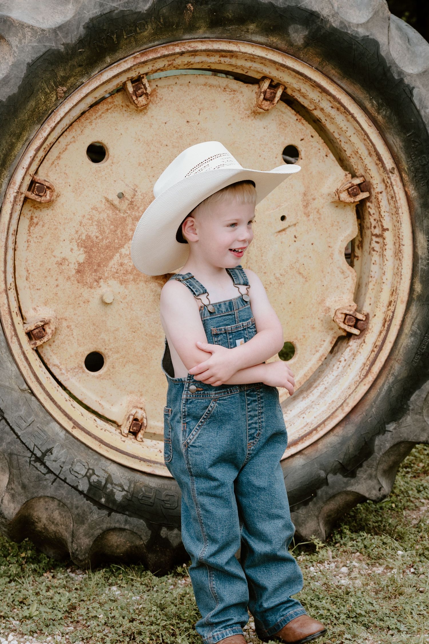A toddler in denim overalls and white cowboy hat standing next to a large tractor tire.