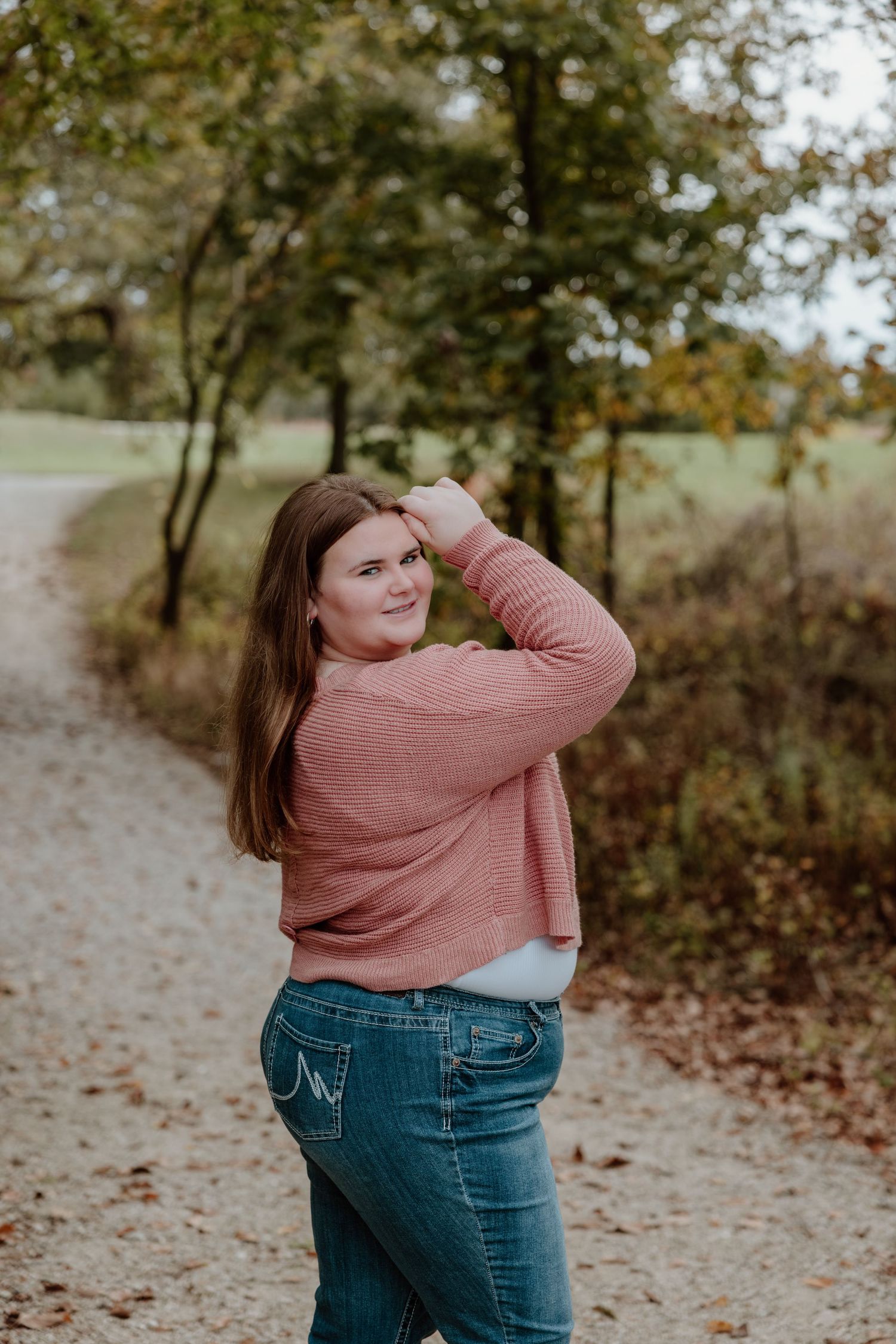 A figure in a pink sweater and jeans poses playfully in a wooded area with fallen autumn leaves.