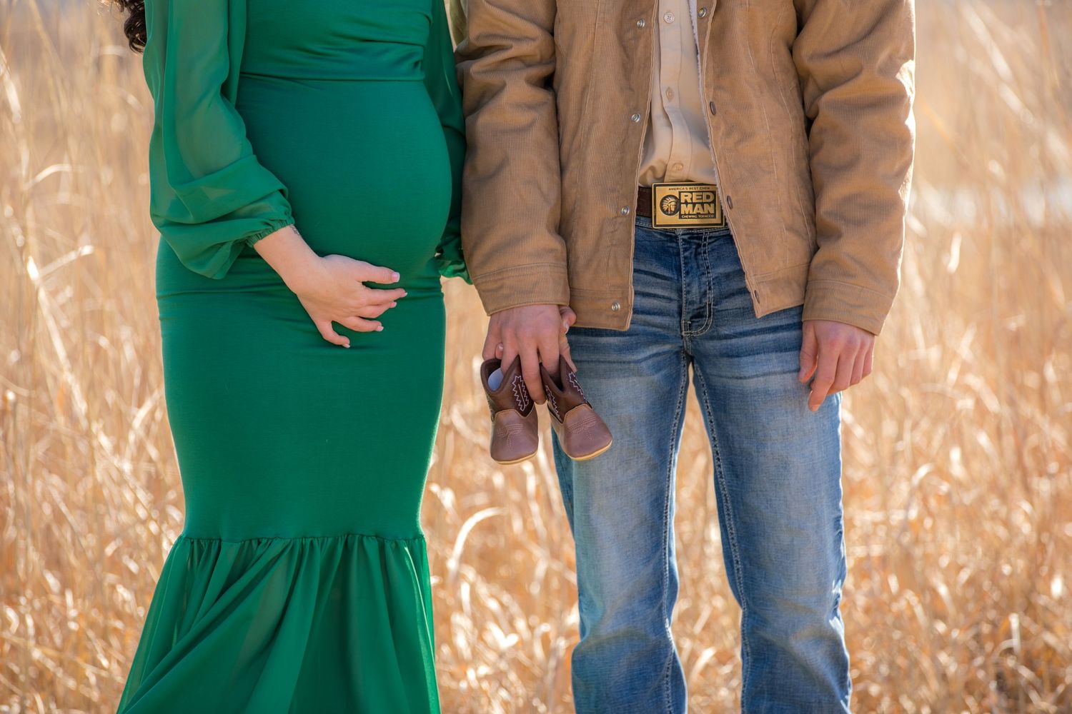A couple stands together in a field showing their maternity style with a green dress and casual denim.