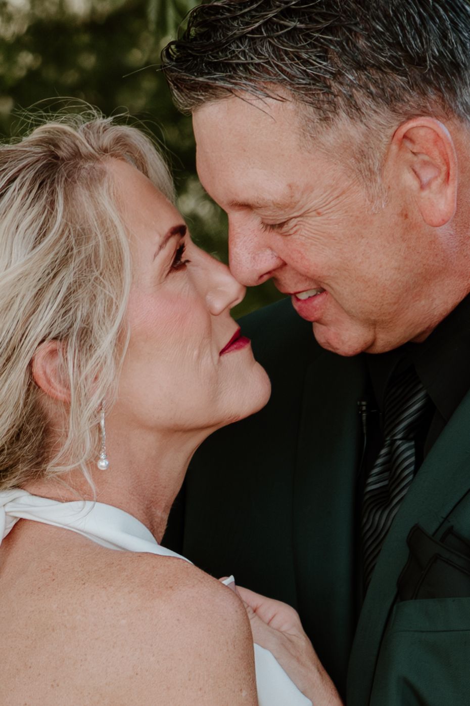 A mature couple in wedding attire share a romantic nose-to-nose moment, smiling tenderly at each other outdoors.