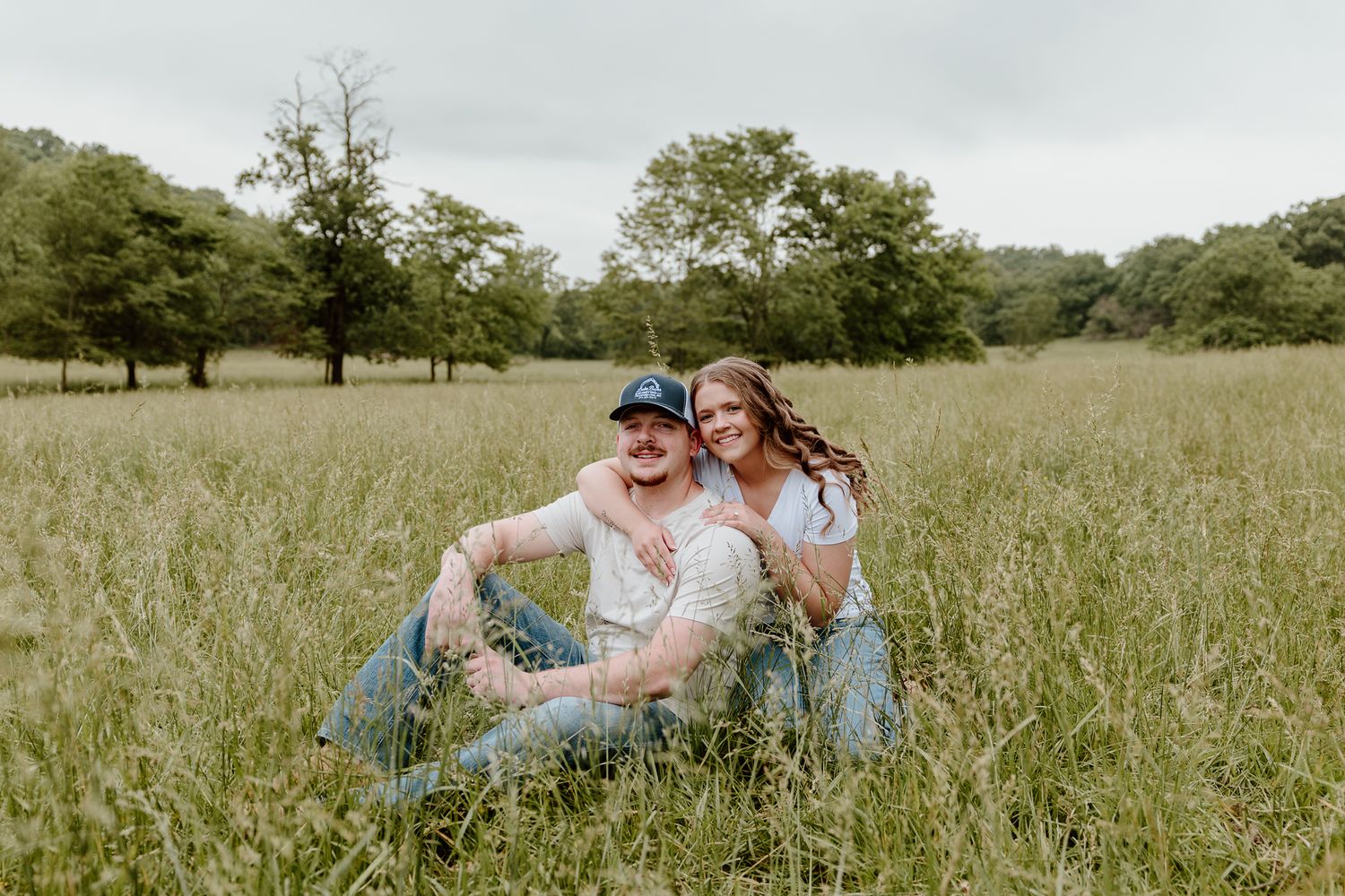 Couple sitting together in a lush green meadow, smiling, with trees in the background on a cloudy day.