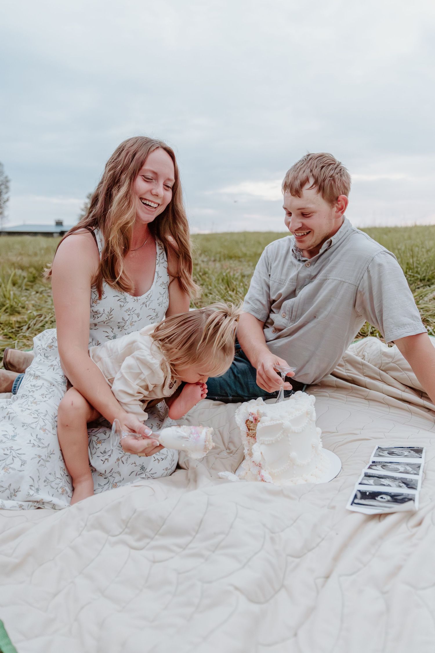 A couple lies on a white blanket in a grassy field while looking at photos together on a cloudy day.