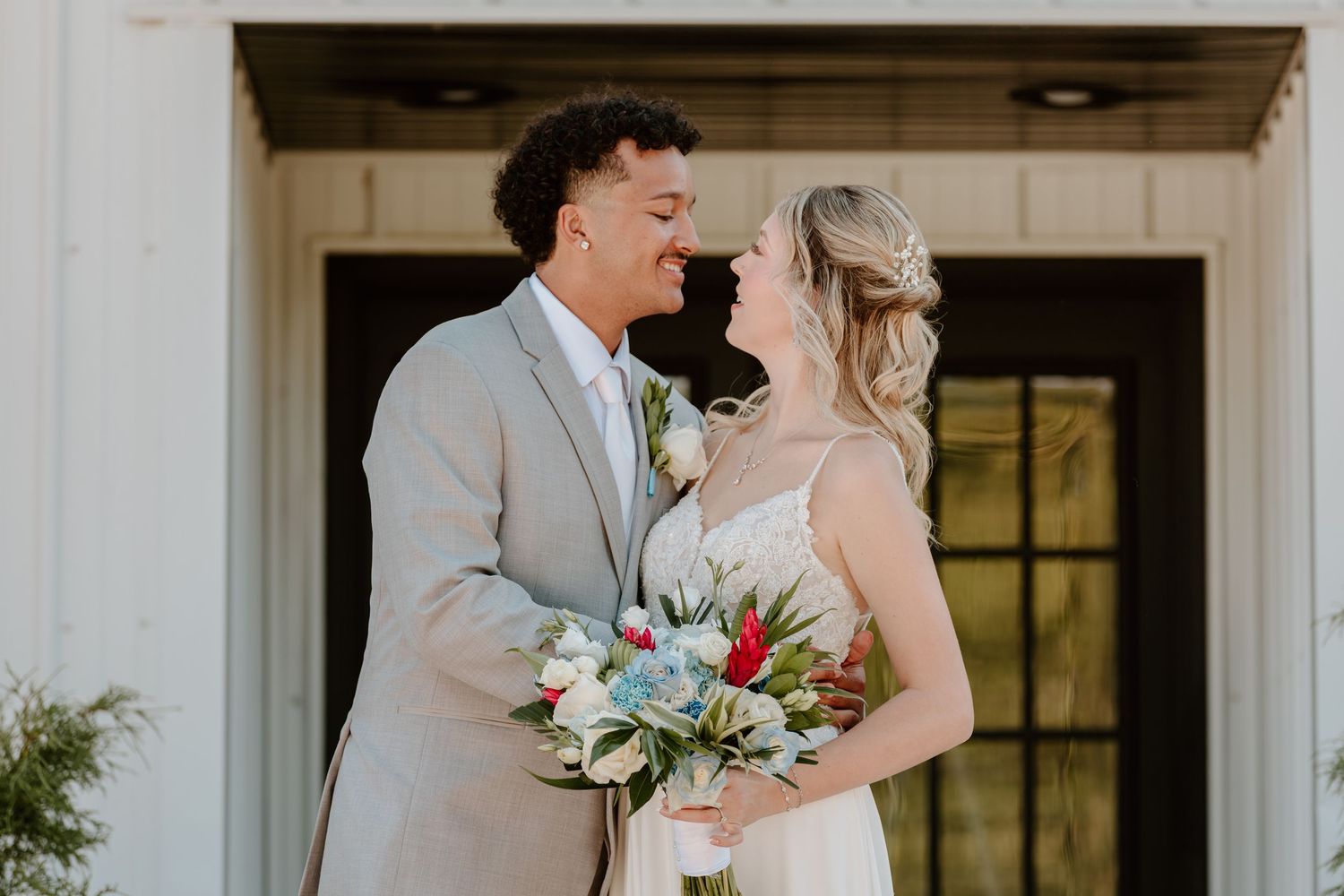 Bride and groom smiling at each other, bride holding a colorful bouquet, standing outside a white building.