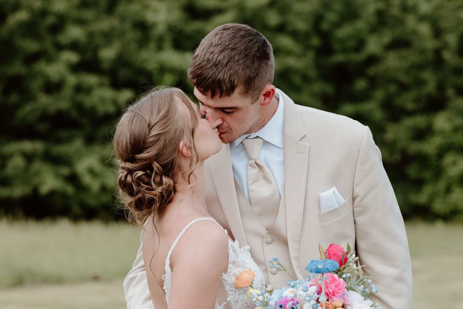 A bride and groom share a romantic kiss outdoors, with the groom wearing a beige suit and bride holding colorful flowers.