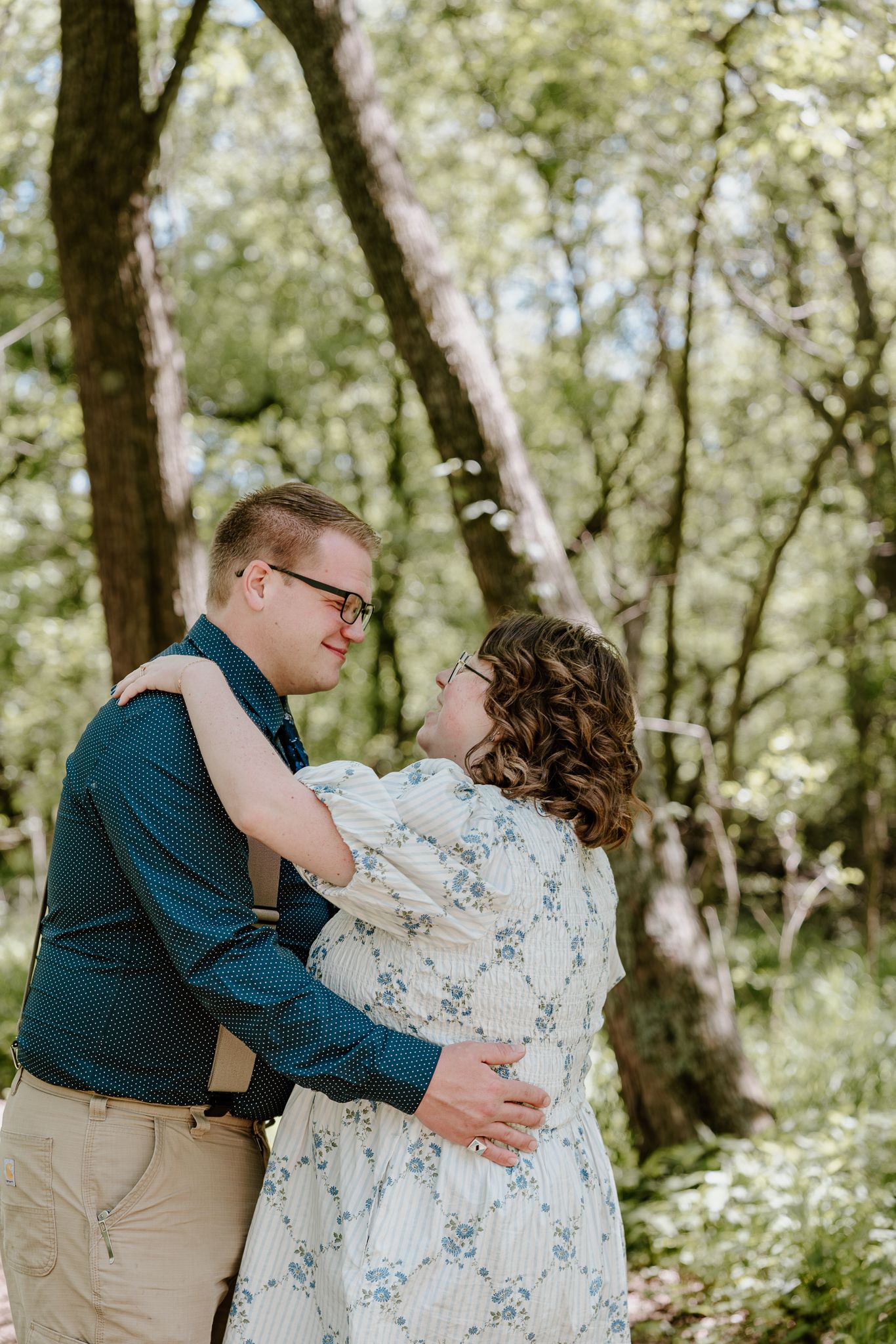 Couple embracing and gazing at each other in a lush green forest setting, woman in floral dress, man in teal shirt.