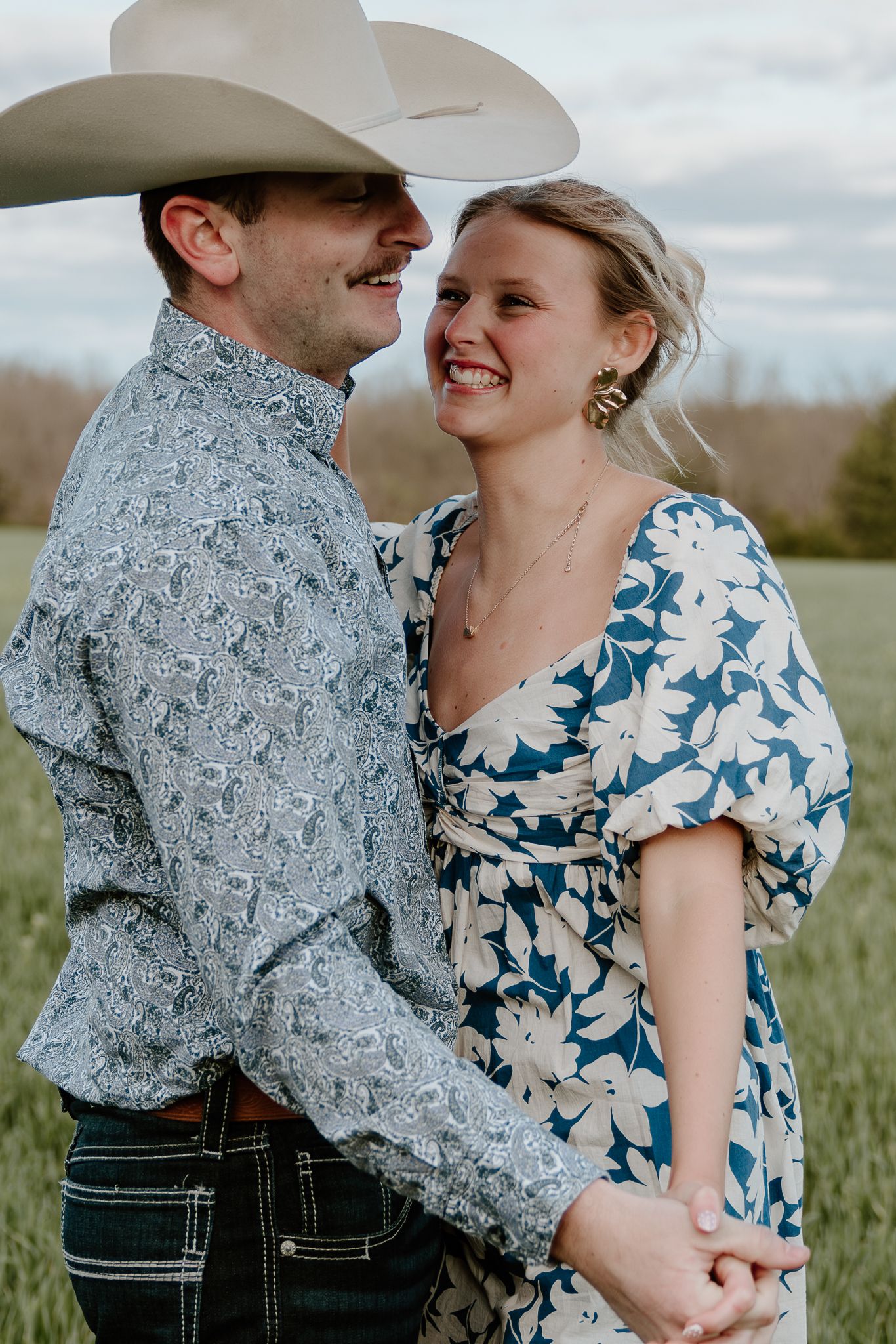 A smiling couple in a field, man in cowboy hat and paisley shirt, woman in blue floral dress, holding hands.