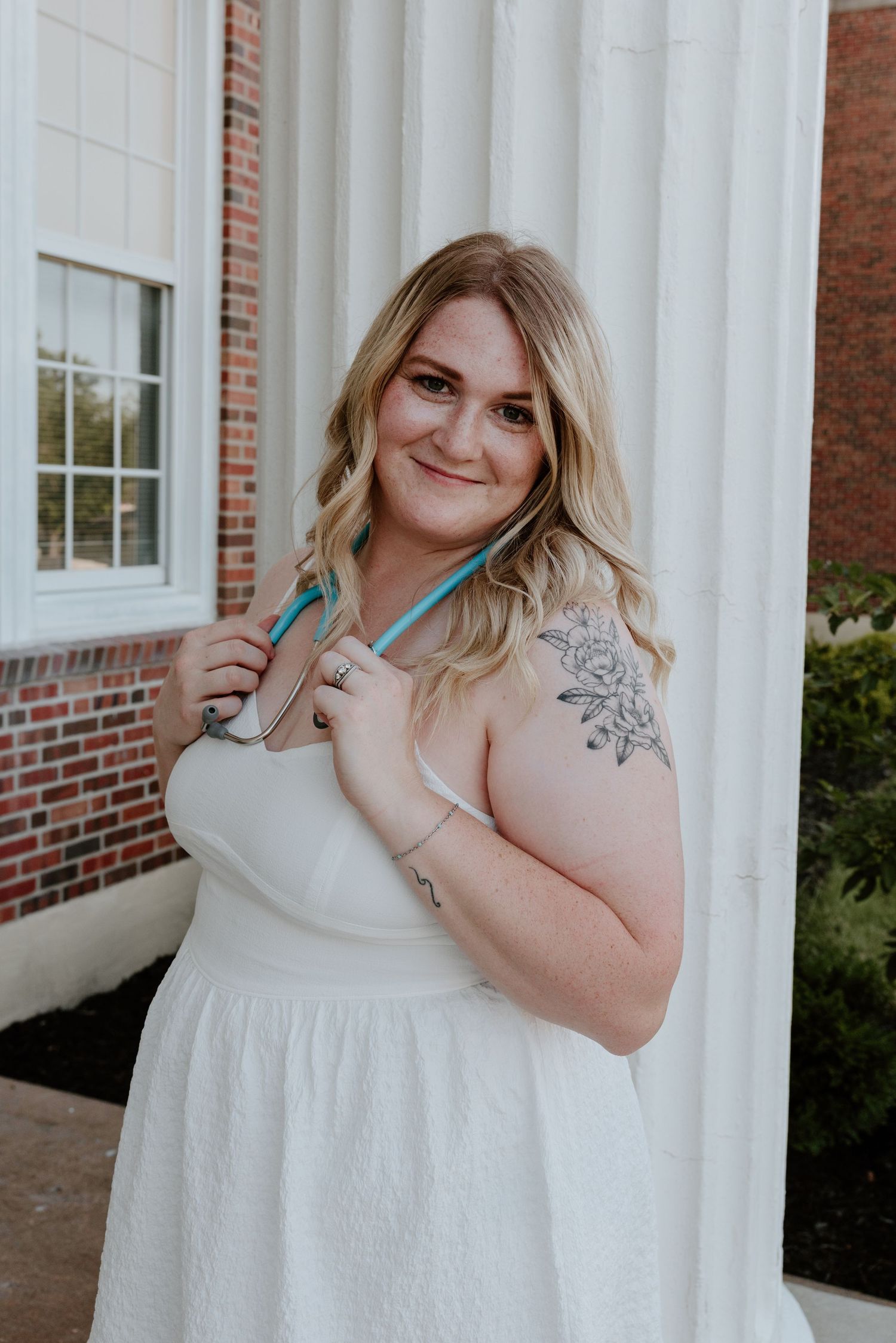 A person in a white dress poses near white columns and brick building, showing a detailed shoulder tattoo.