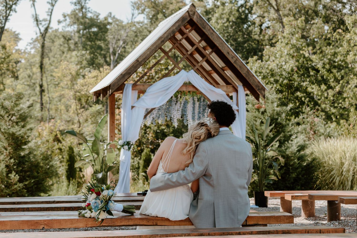 Bride and groom sit together under wooden arch gazebo at outdoor wedding ceremony.