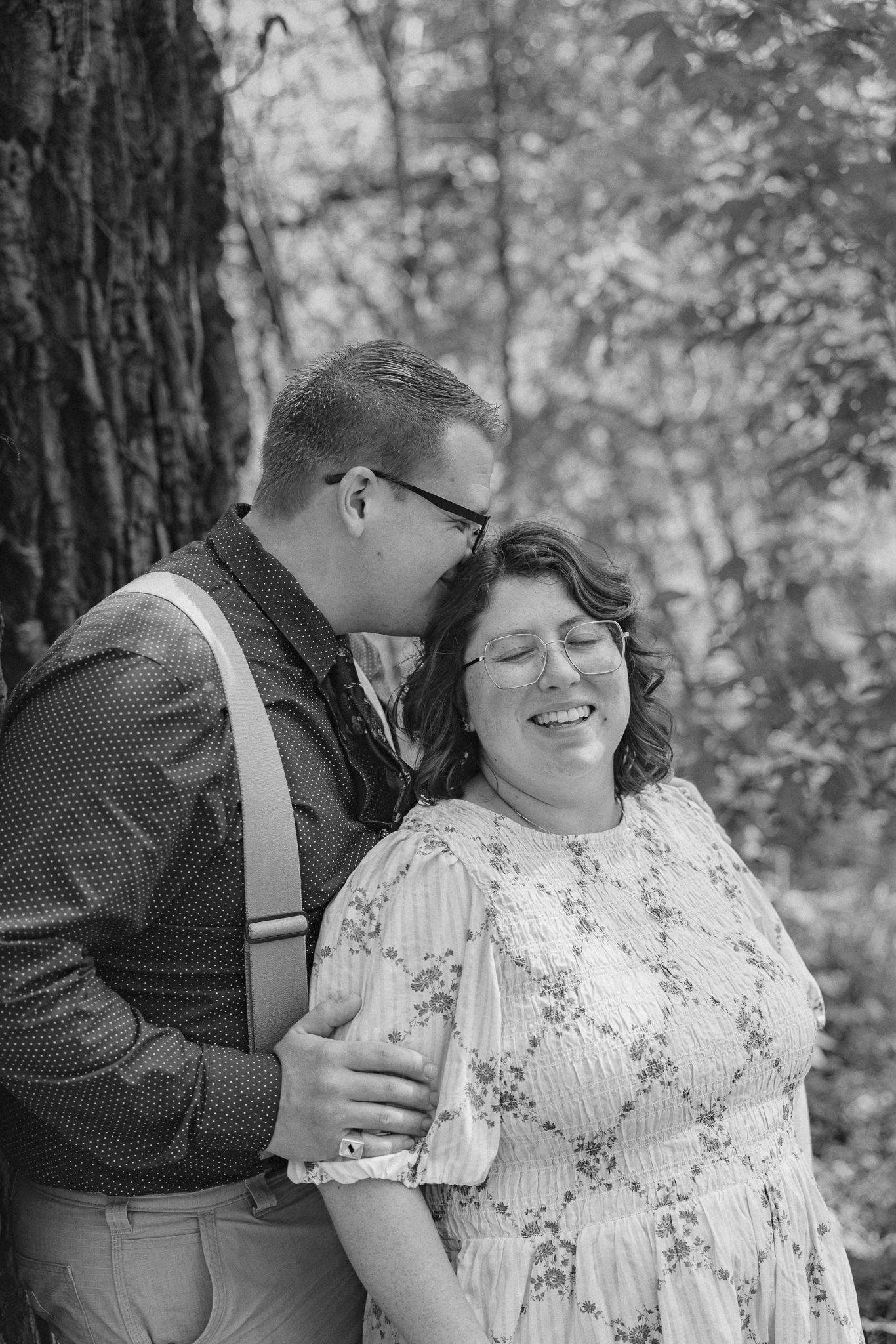 Black and white photo of a couple in a forest, man kissing woman on cheek as she smiles joyfully.