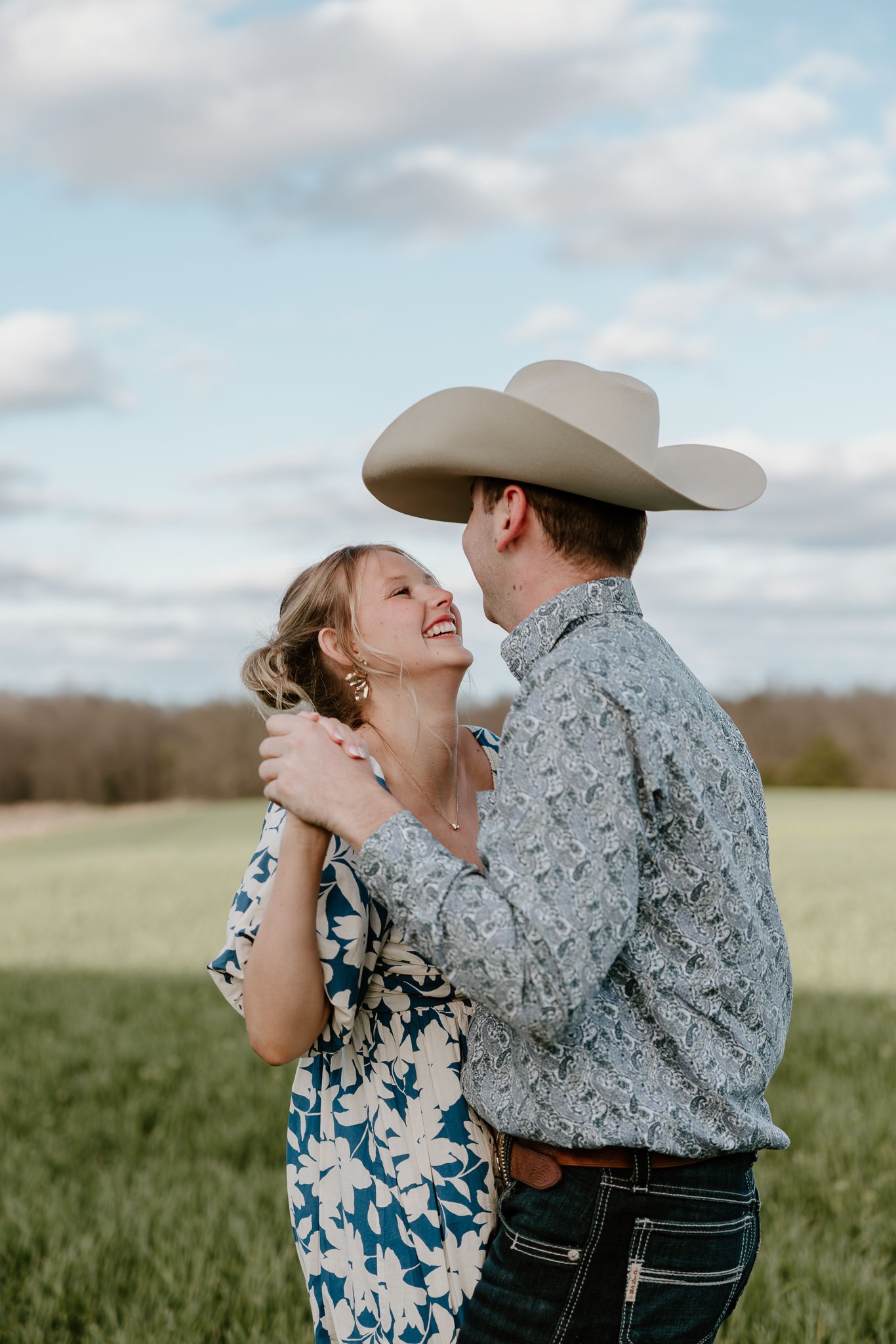 A couple shares a joyful embrace in a green field, the man wearing a cowboy hat and the woman in a floral dress.