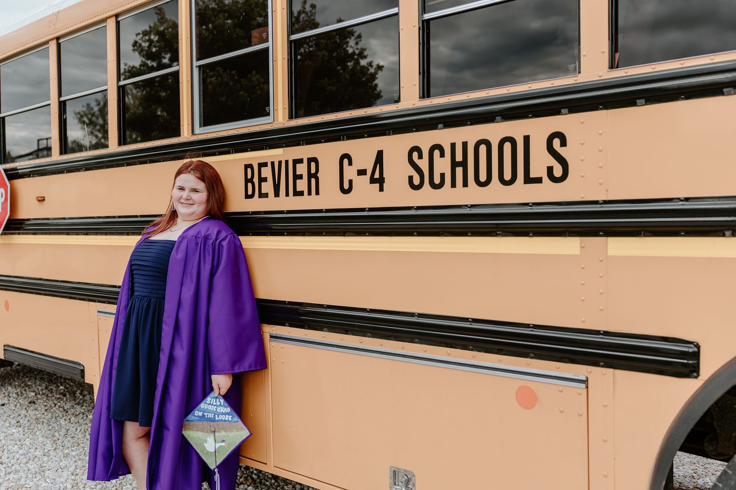 A graduate in purple cap and gown poses proudly beside a Bevier C-4 Schools yellow school bus.