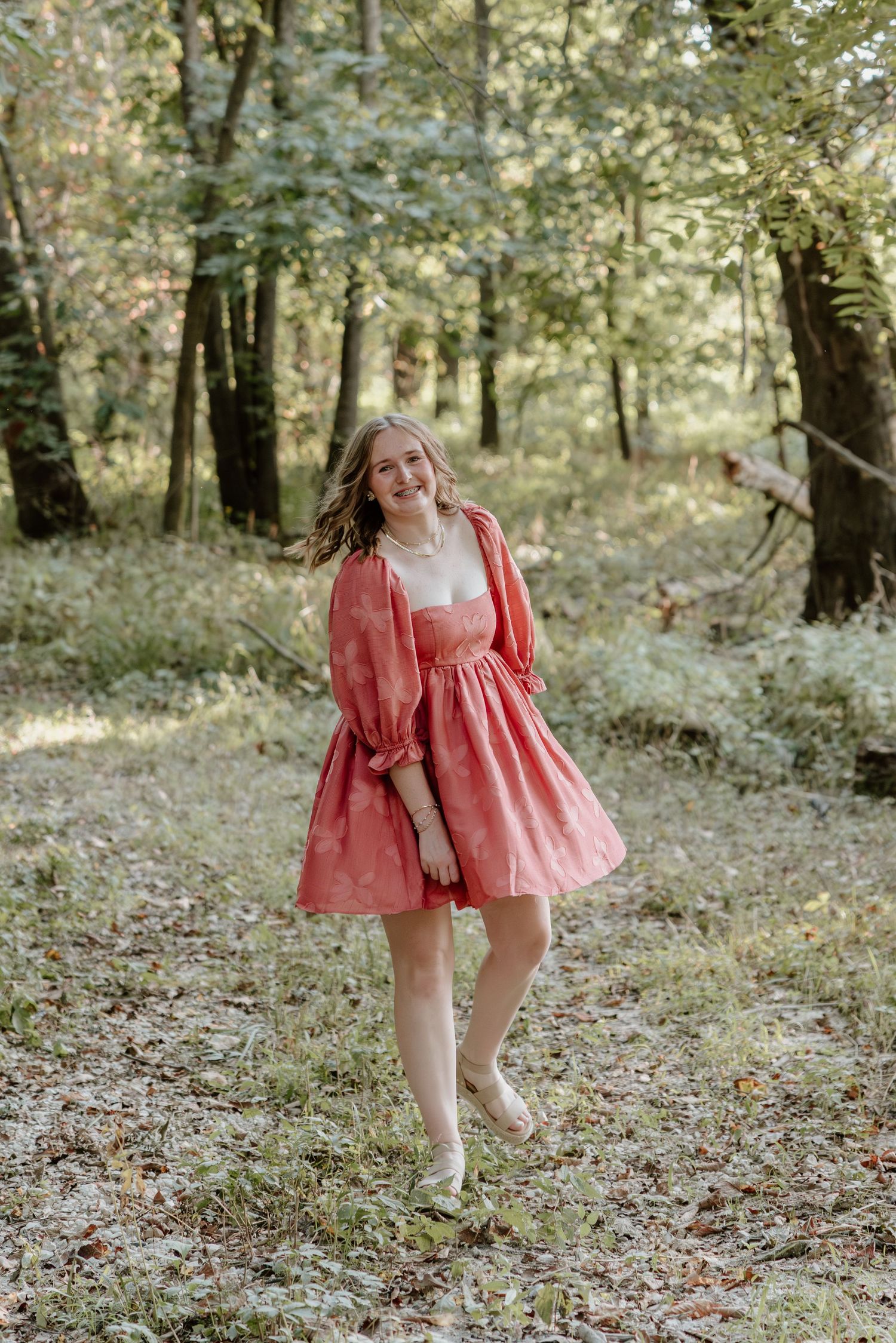 A person in a flowing coral dress twirls and dances in a sunlit wooded area covered with fallen leaves.