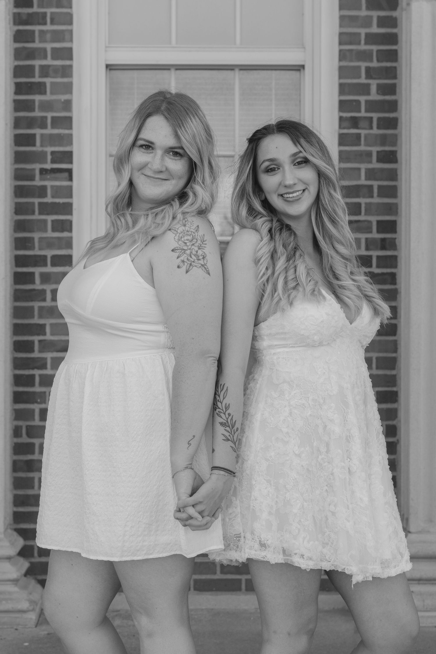 Group wearing white lace dresses poses together against brick wall in casual summer photoshoot.