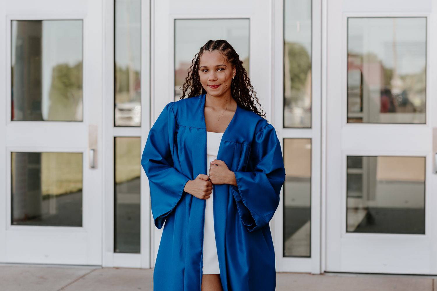 Young woman in blue graduation gown smiling outside school building on graduation day.