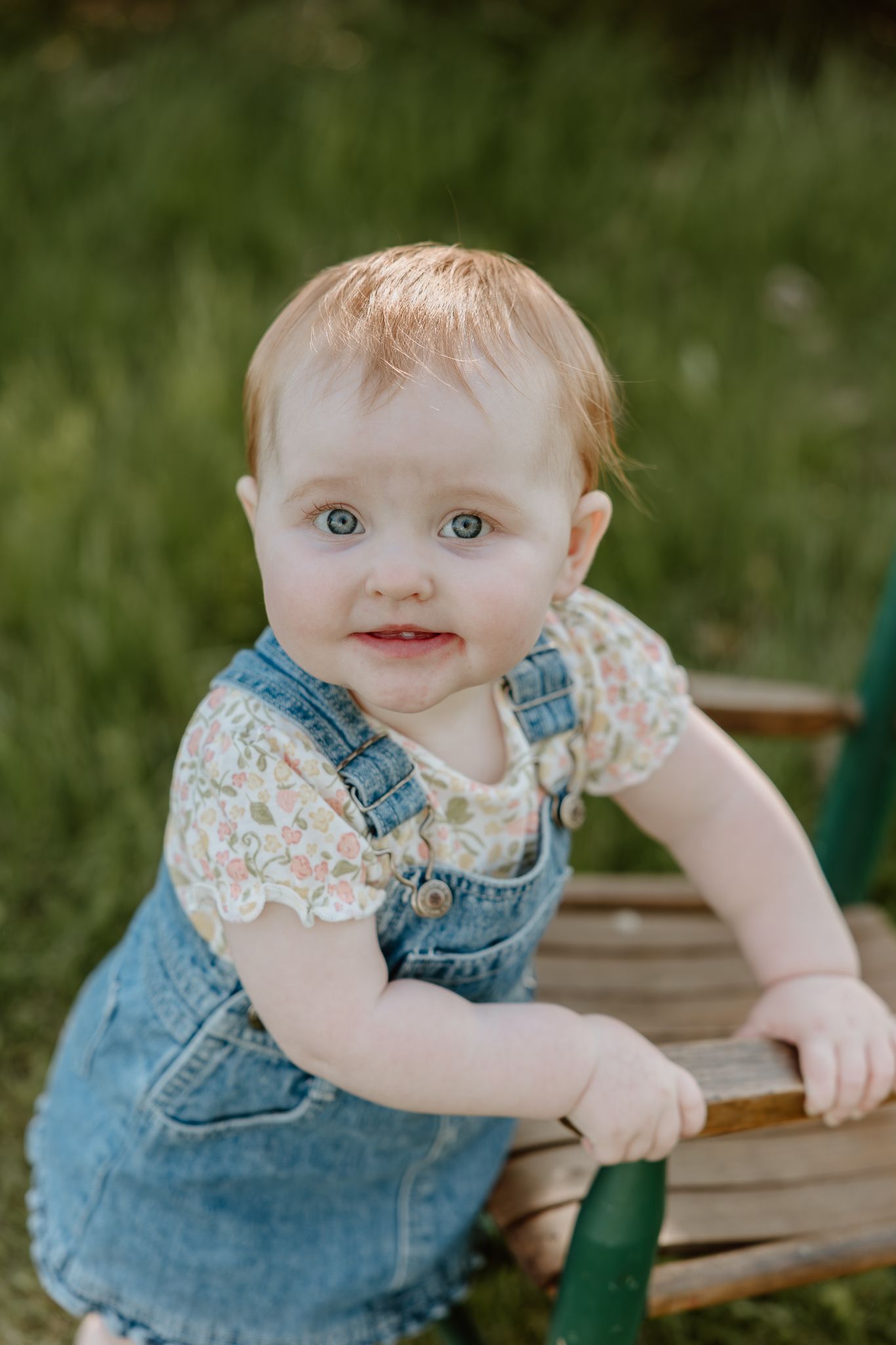 Adorable baby girl in denim overalls and floral shirt smiling outdoors with green grass background.