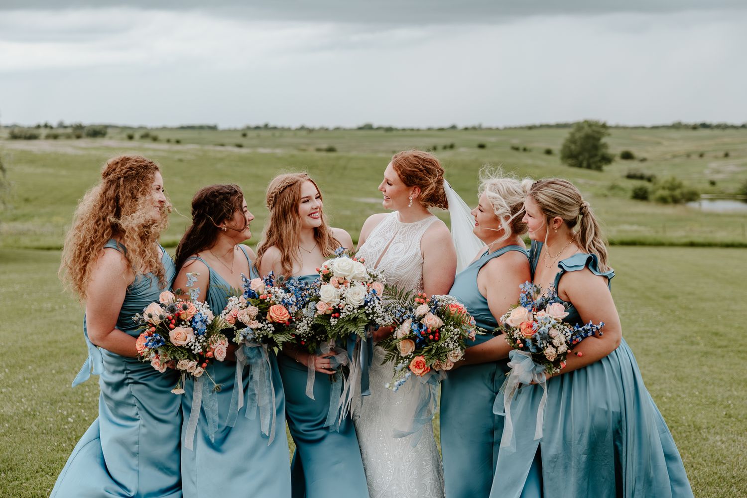 Bride and five bridesmaids in blue dresses laughing together outdoors holding colorful floral bouquets.