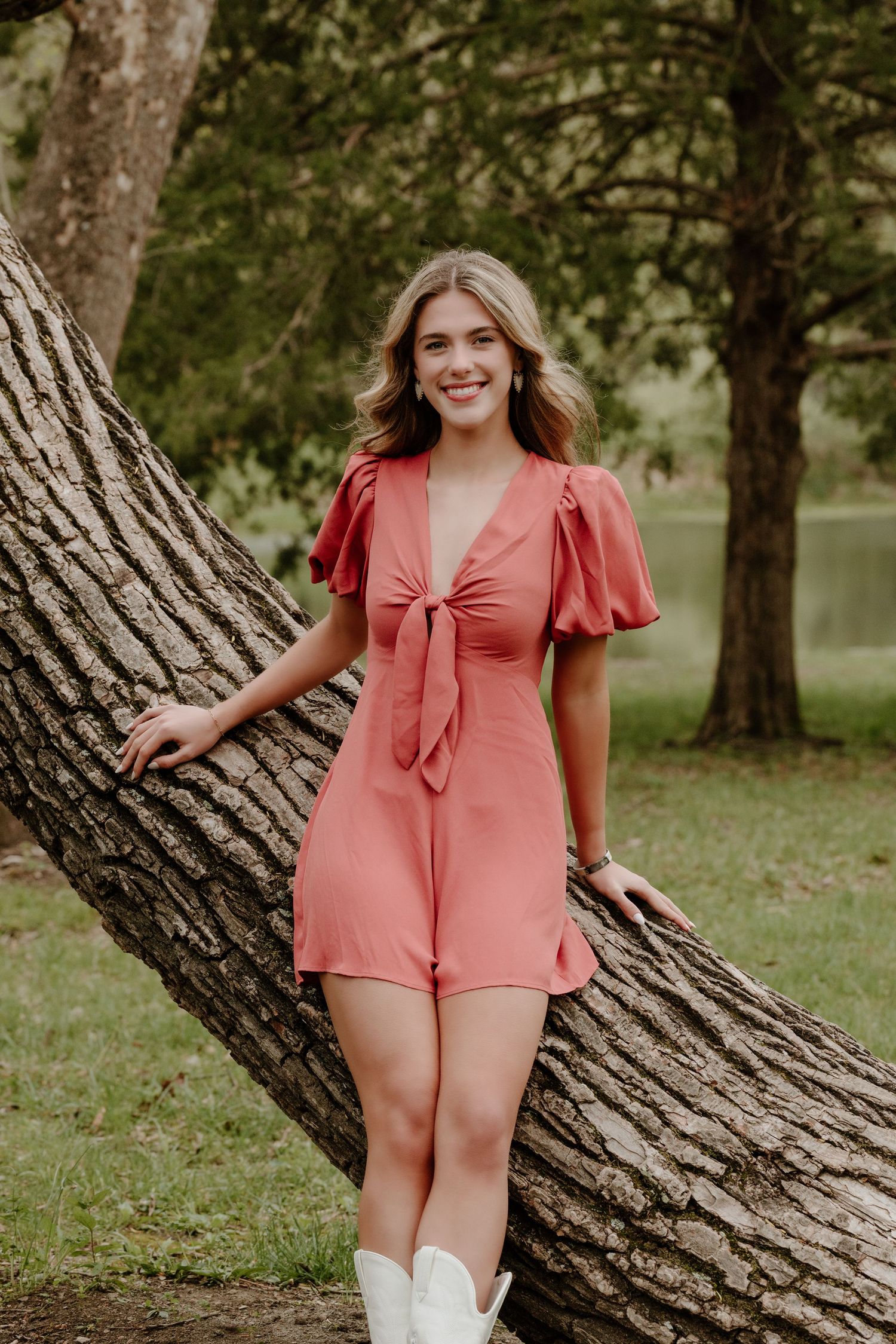 A woman in a coral-colored romper with puff sleeves and white boots poses casually against a large tree trunk near a lake.