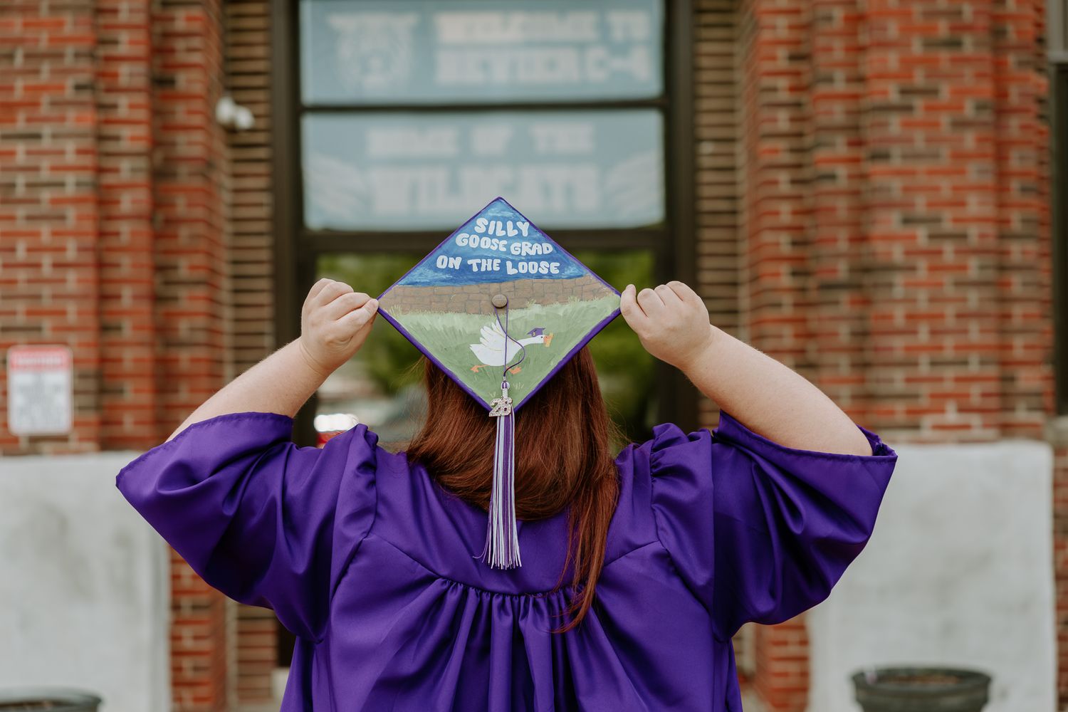 Graduate in purple gown holds decorated cap showing Bills stadium design at outdoor graduation ceremony.