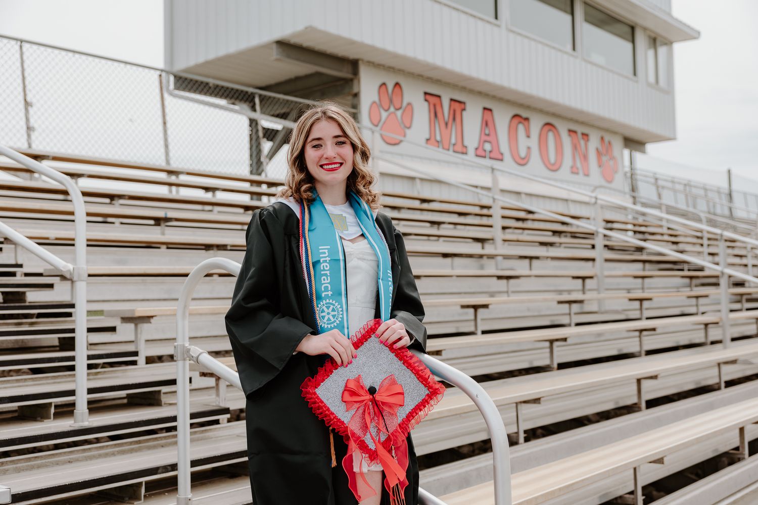 Smiling graduate in black gown and blue sash holds decorated cap at Macons stadium bleachers.