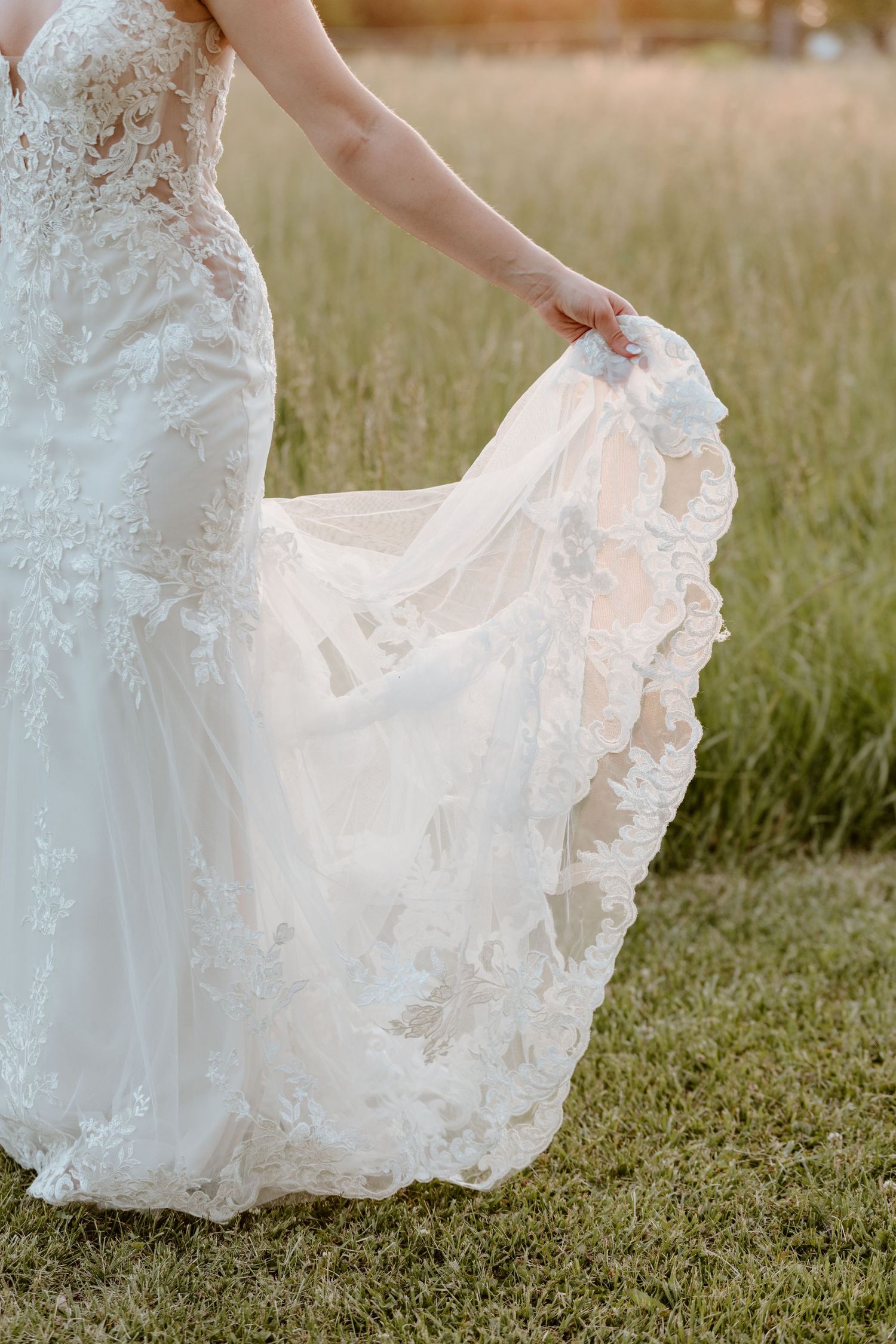 Bride lifting lace wedding dress train in a grassy outdoor field during golden hour.