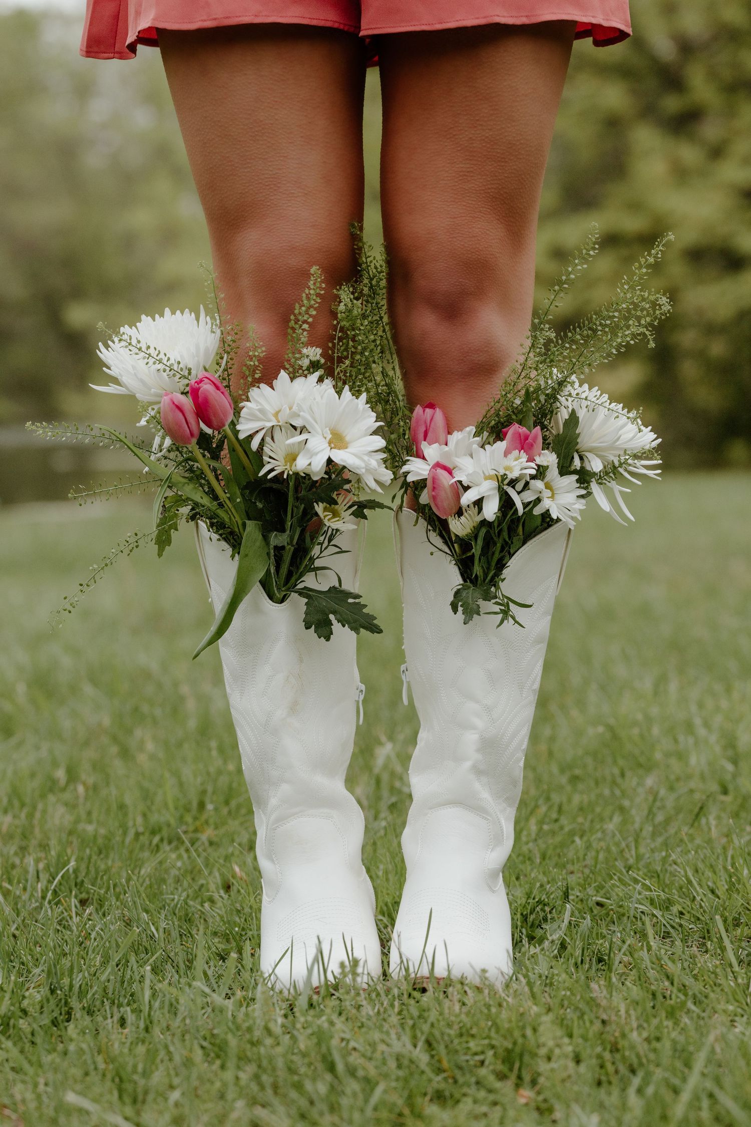 White rubber rain boots filled with fresh daisies and pink flowers stand on green grass with a red umbrella in view.