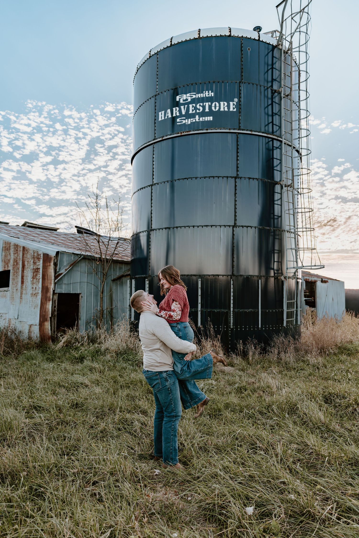 A couple shares an intimate moment in front of large grain silos at sunset.