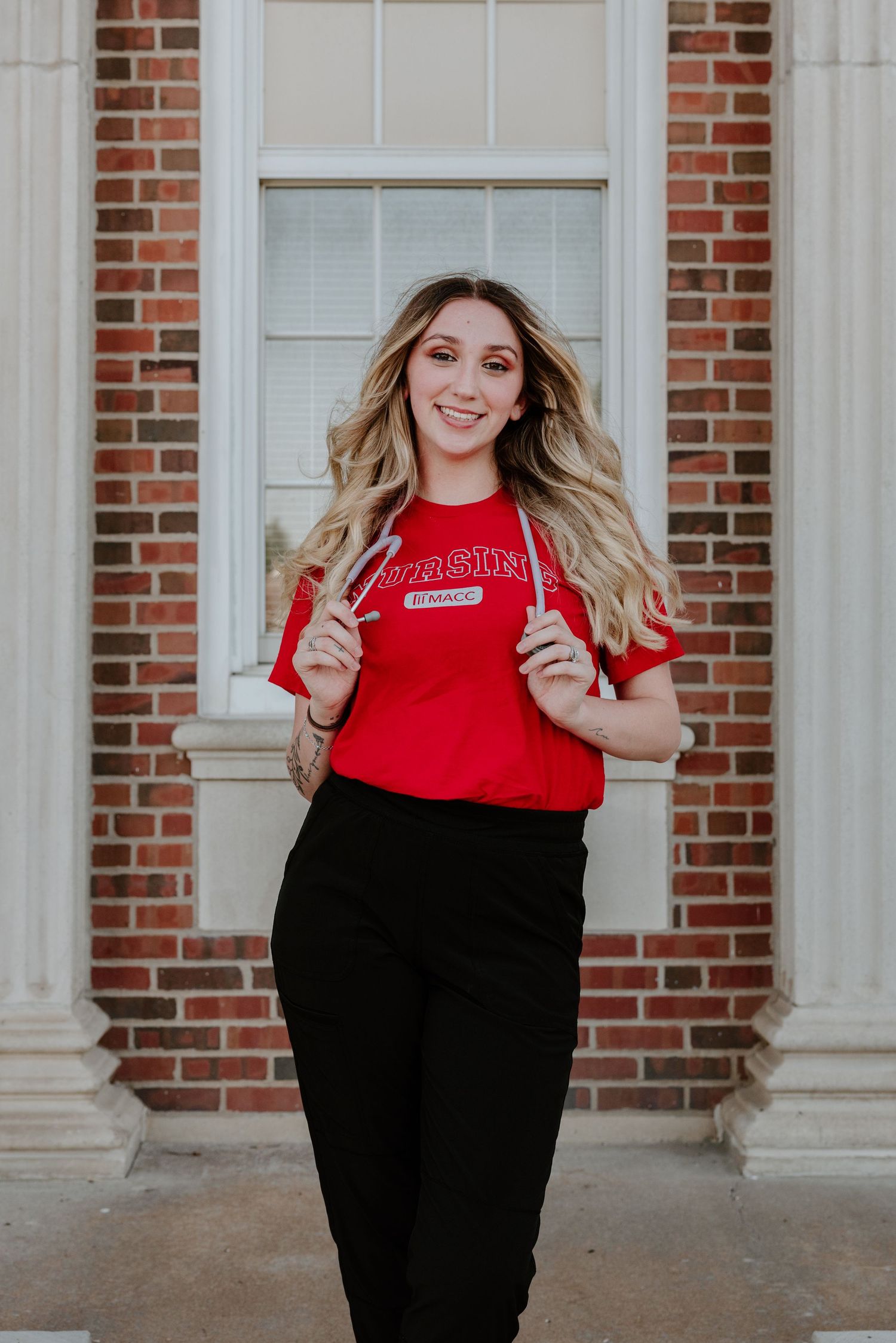 A person in a red collegiate sweater and black pants stands smiling against a brick building with white columns.