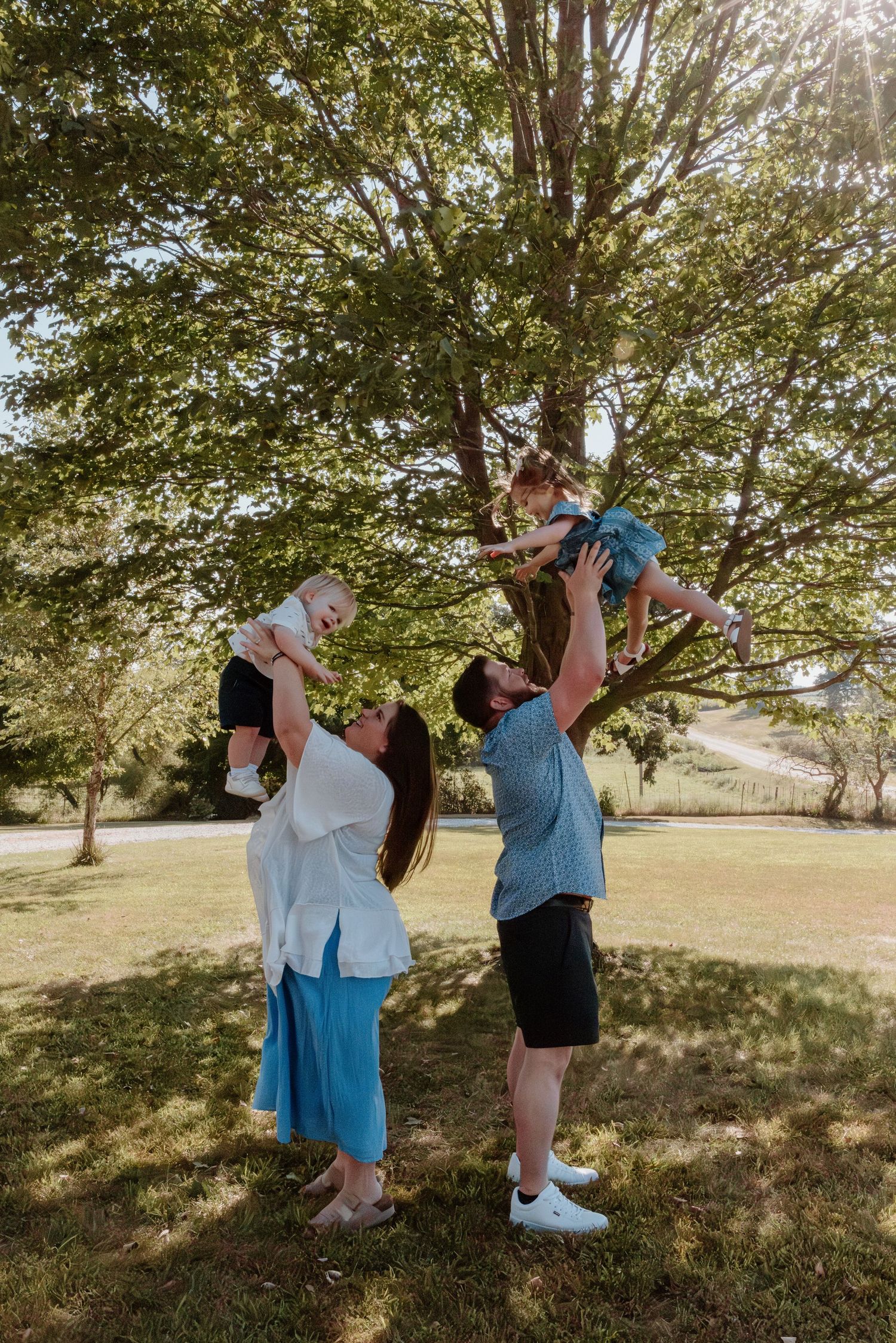 Two people in blue and black outfits dance playfully under trees in a sunny outdoor setting.