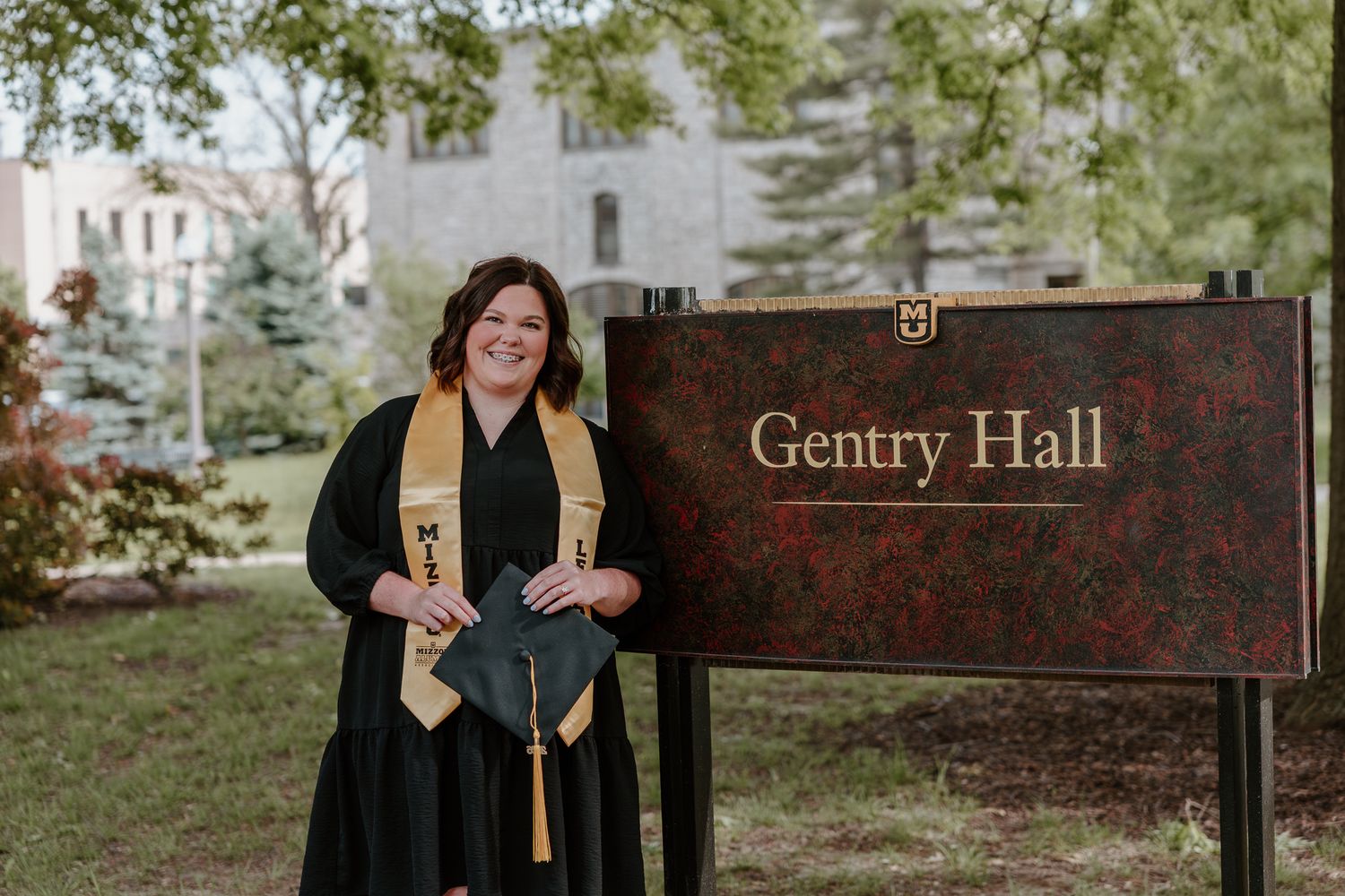 Graduate in cap and gown smiling beside Gentry Hall sign during outdoor graduation photo session.