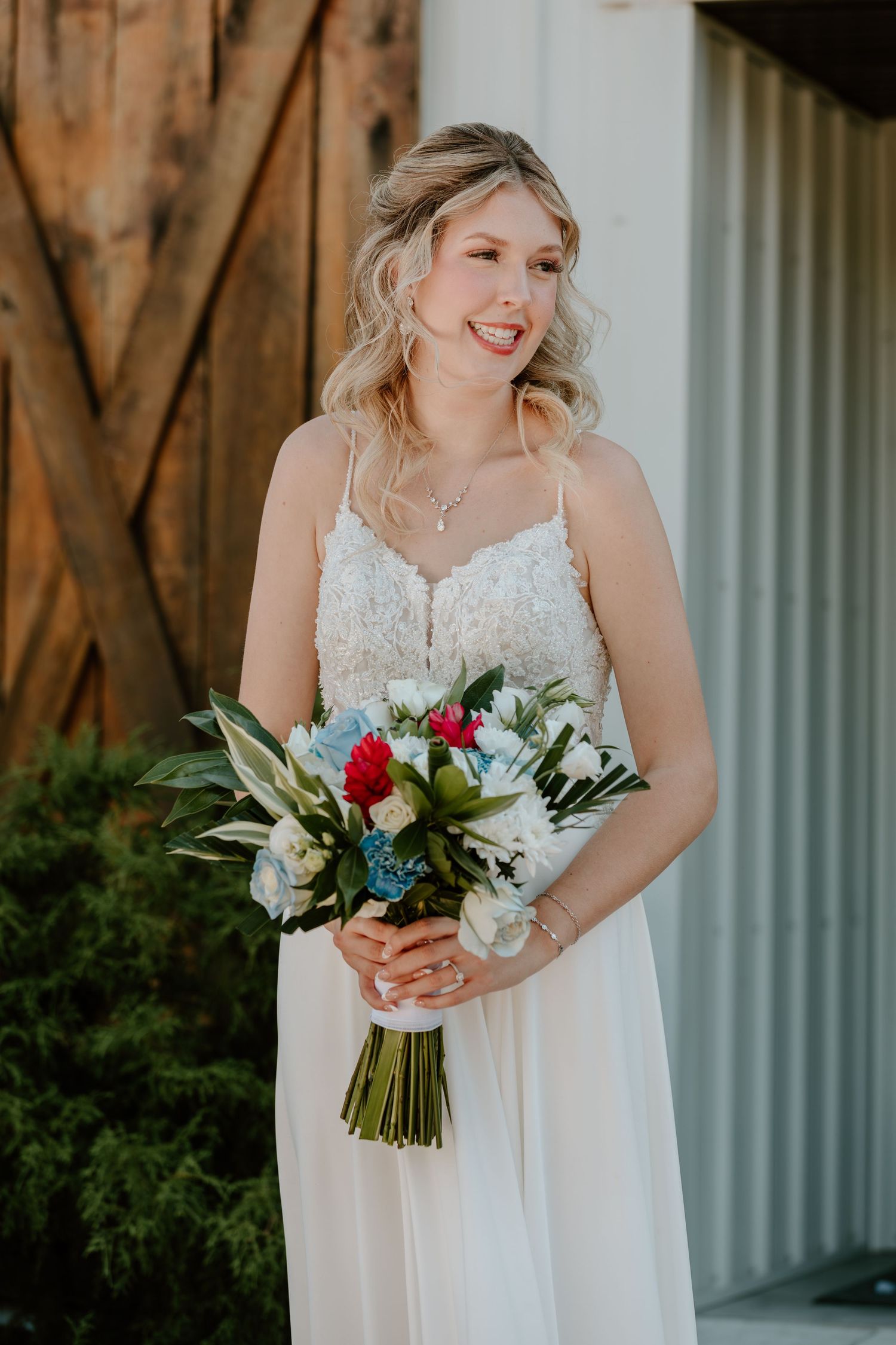 Smiling bride in lace spaghetti strap gown holds colorful red, white, and blue bridal bouquet.