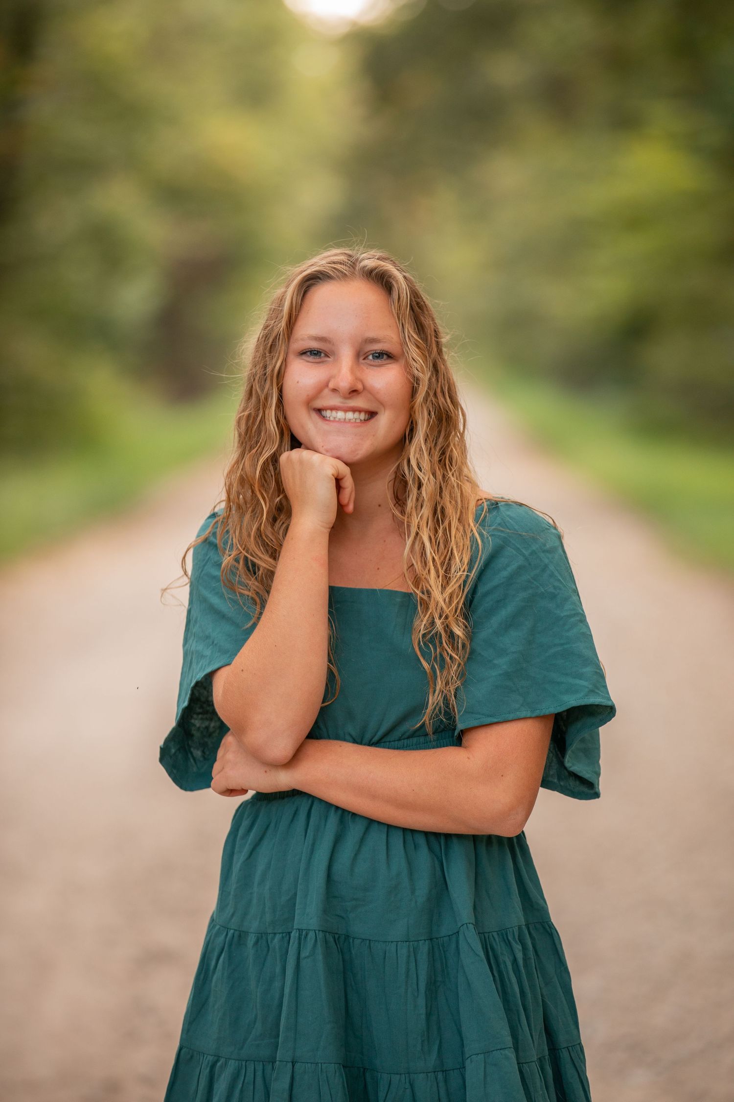 Person in a teal dress smiling on a rural path with bokeh background.
