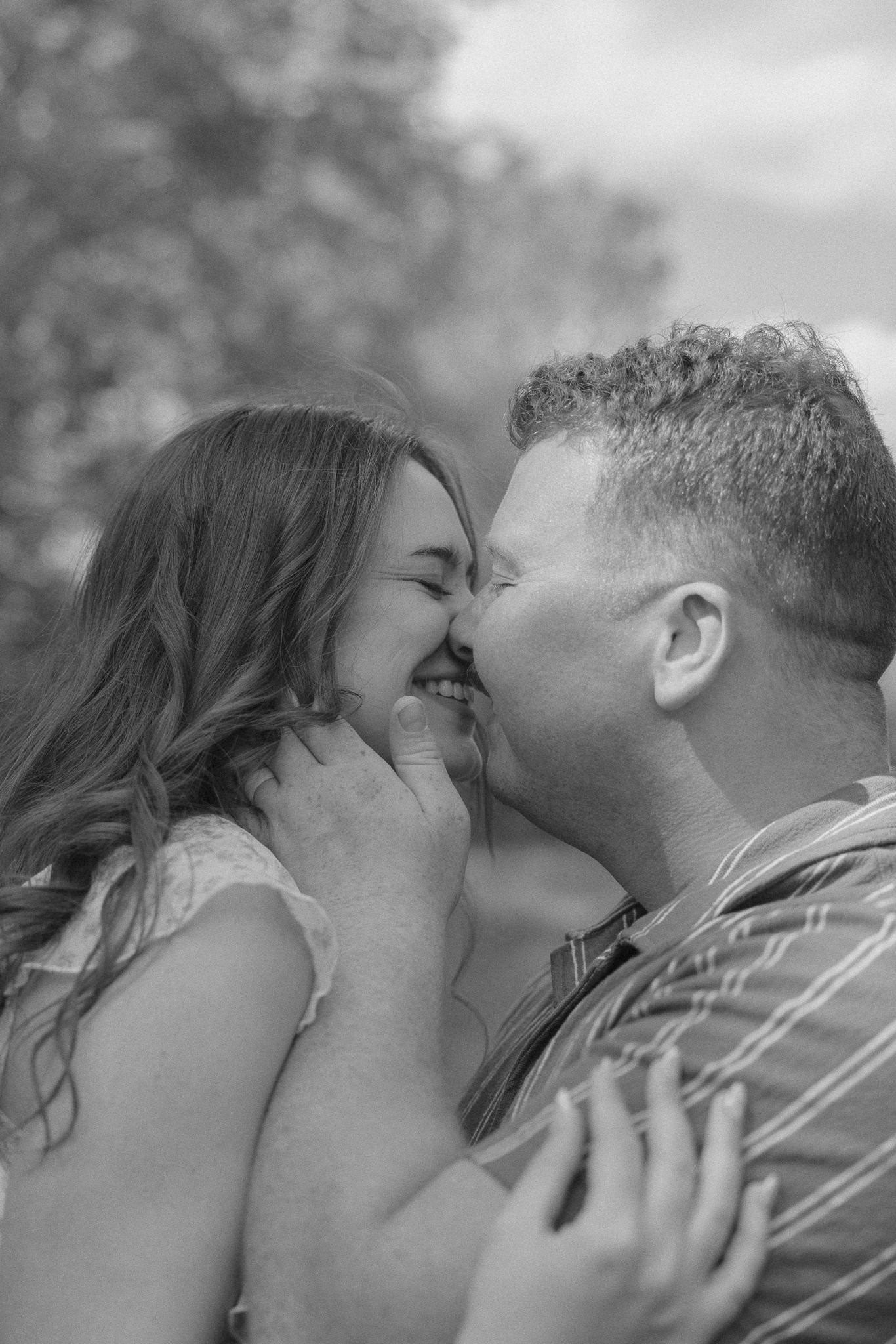 Black and white photo of a couple sharing a tender nose-to-nose moment, smiling and embracing outdoors.