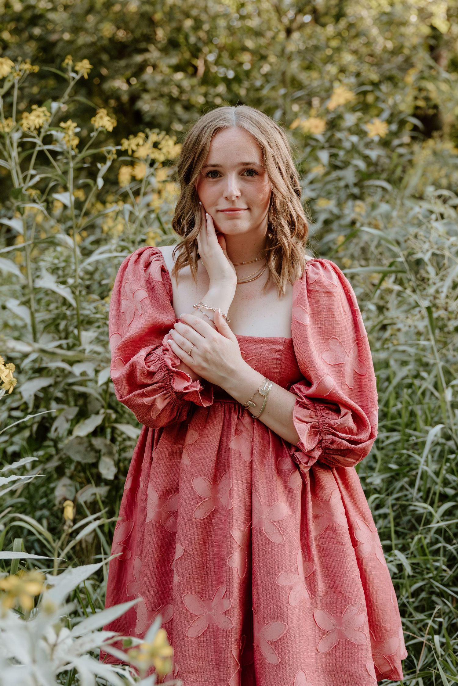 A person wearing a coral pink textured dress poses in a garden setting with natural greenery and flowers.