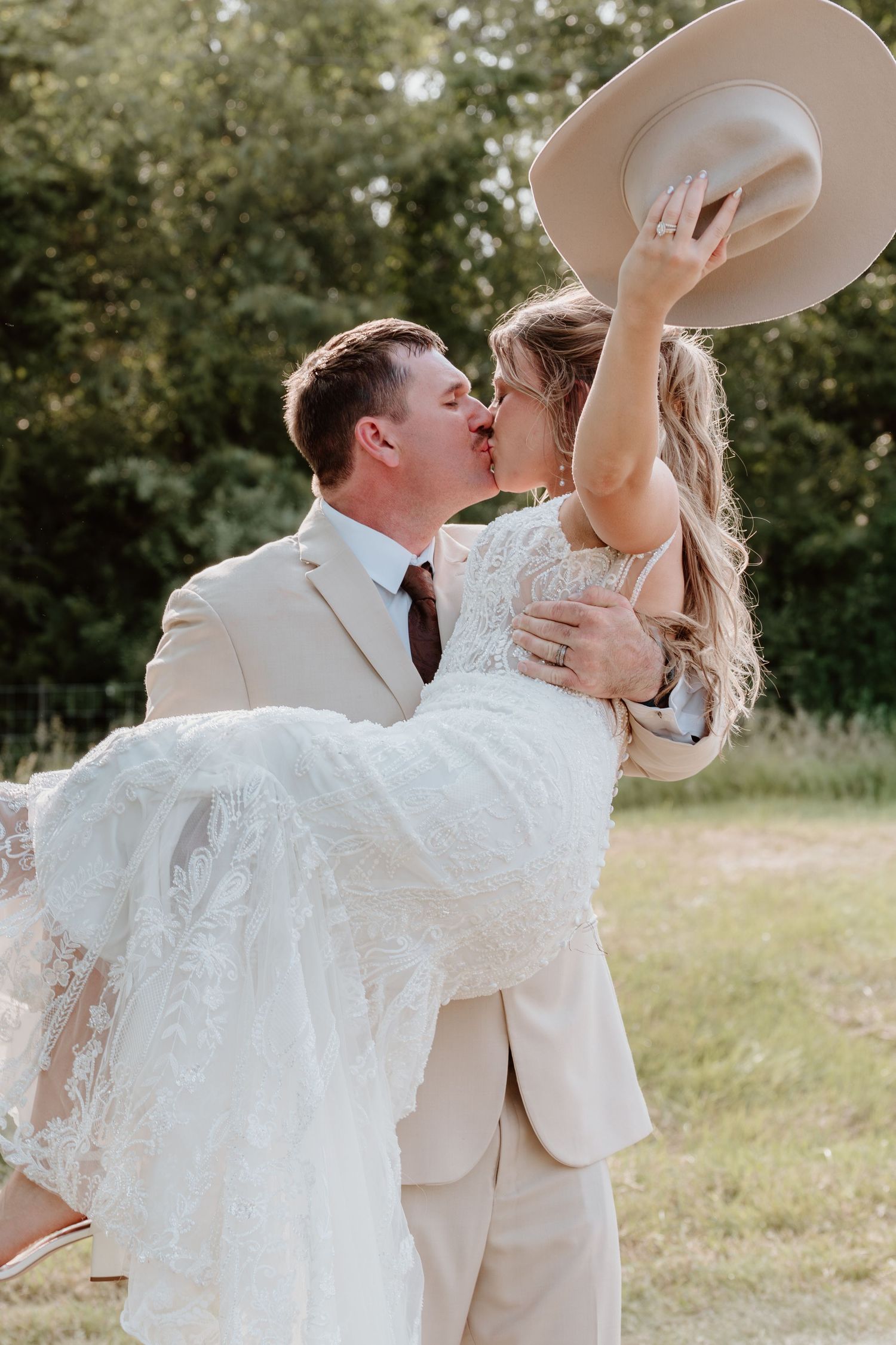 Groom carrying bride while kissing outdoors, bride holding tan cowboy hat in the air.