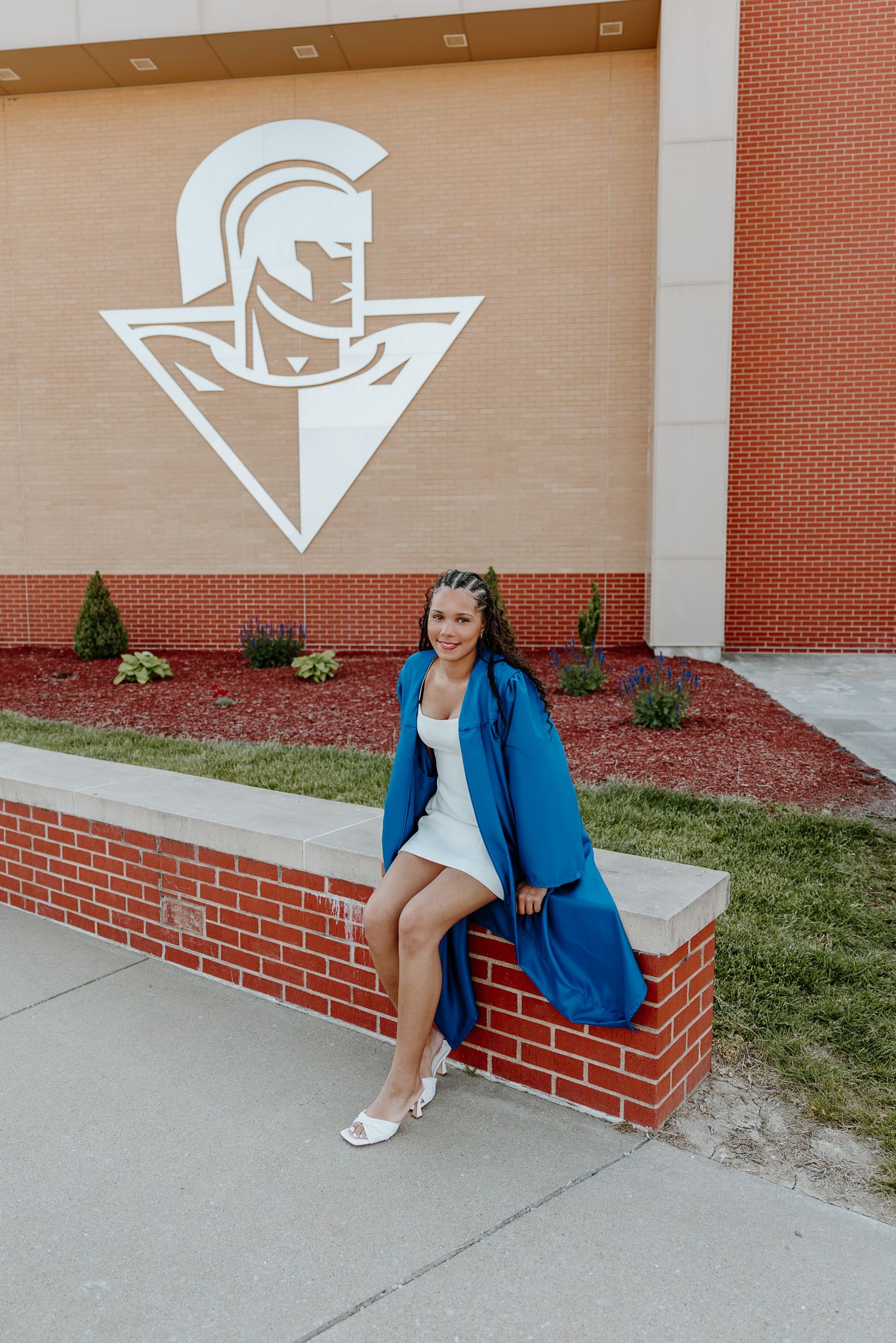 Young woman in blue graduation gown sits on brick wall outside school with Trojan mascot logo.