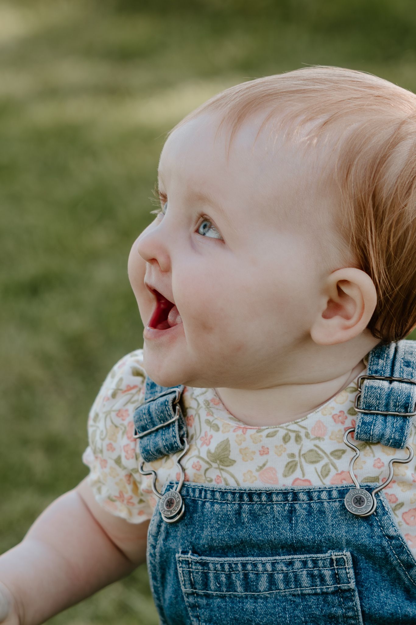 Adorable baby girl in denim overalls and floral shirt smiling outdoors on green grass background.