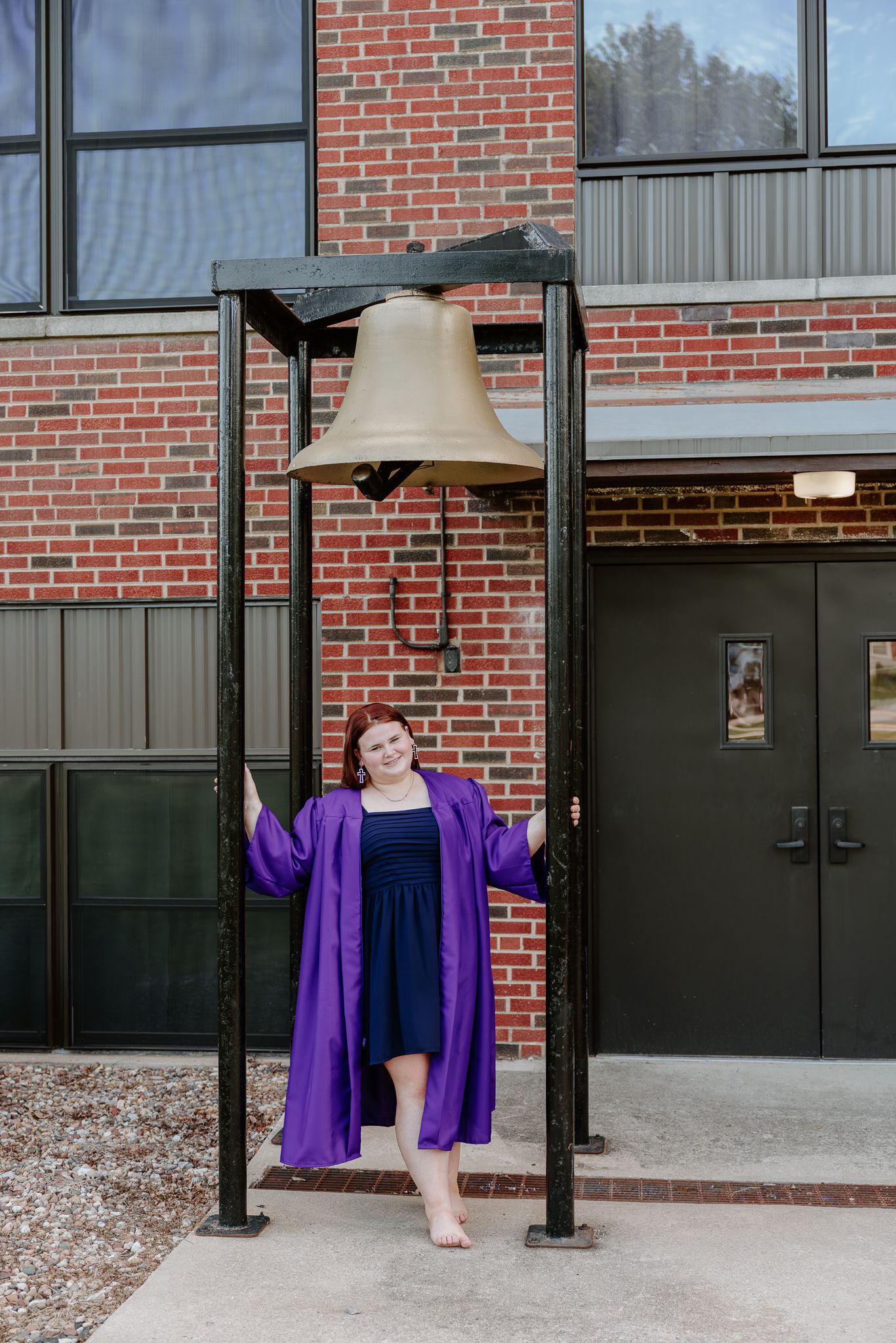 Graduate in purple robe poses barefoot beside a large bronze bell outside a brick building.