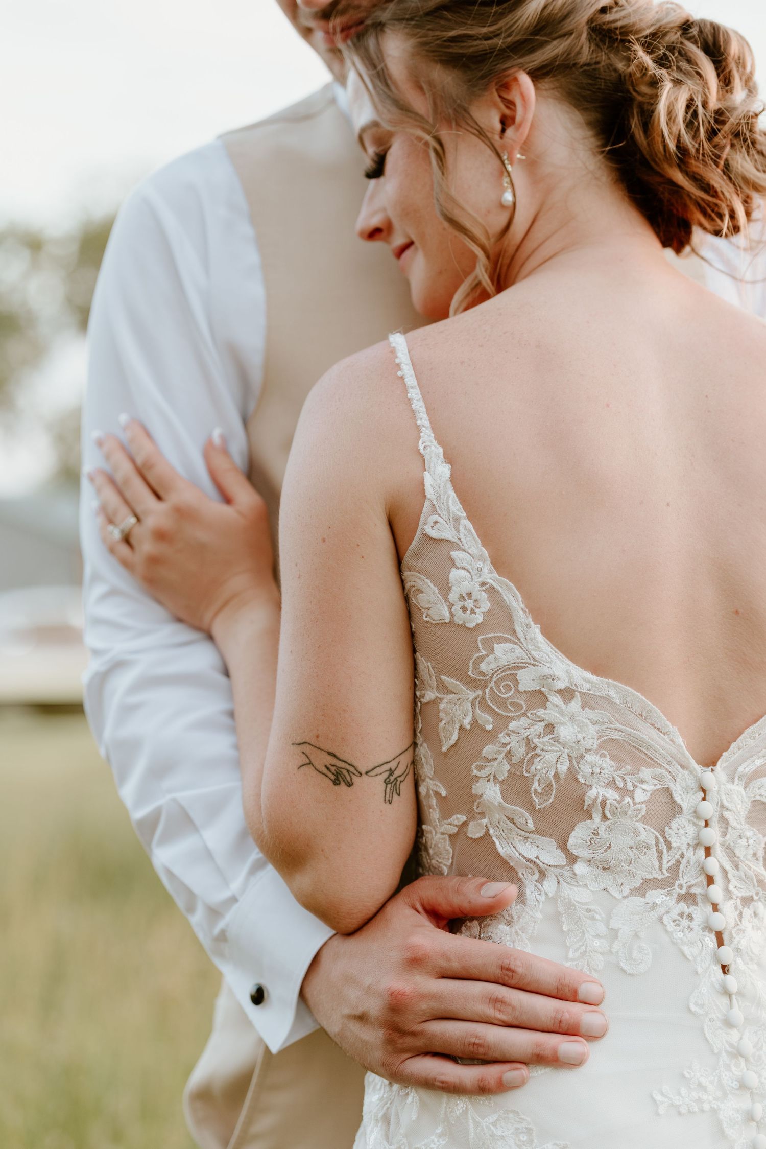 Bride in lace wedding gown with floral tattoo embraces groom in white shirt outdoors.