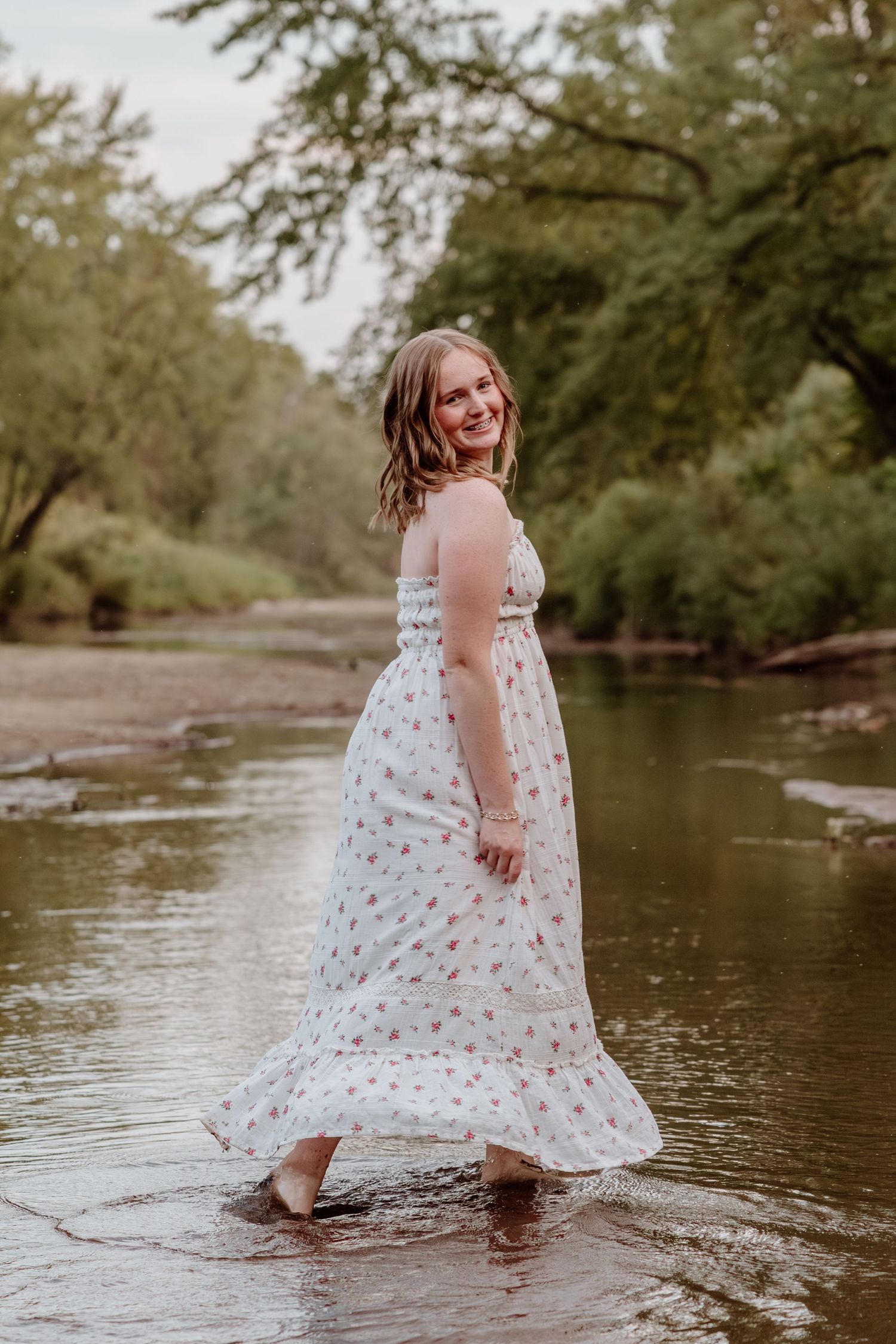 Child in a white floral dress wading through shallow river water on a summer day.