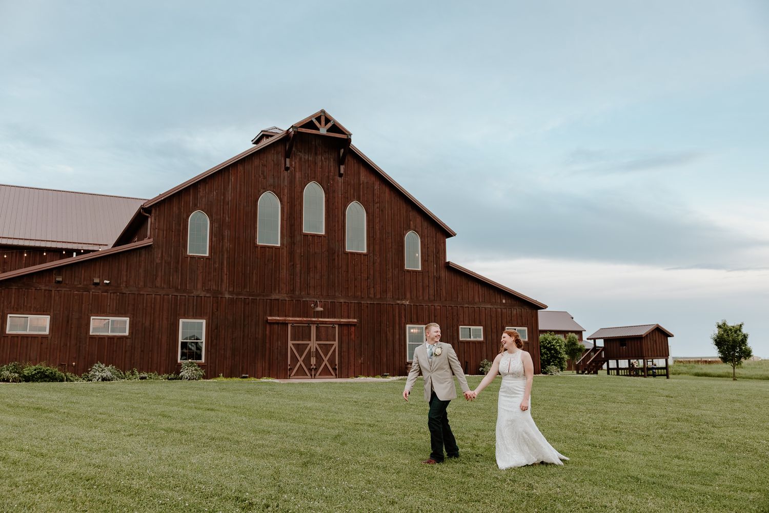 Bride and groom hold hands walking across green lawn in front of a large red barn wedding venue.