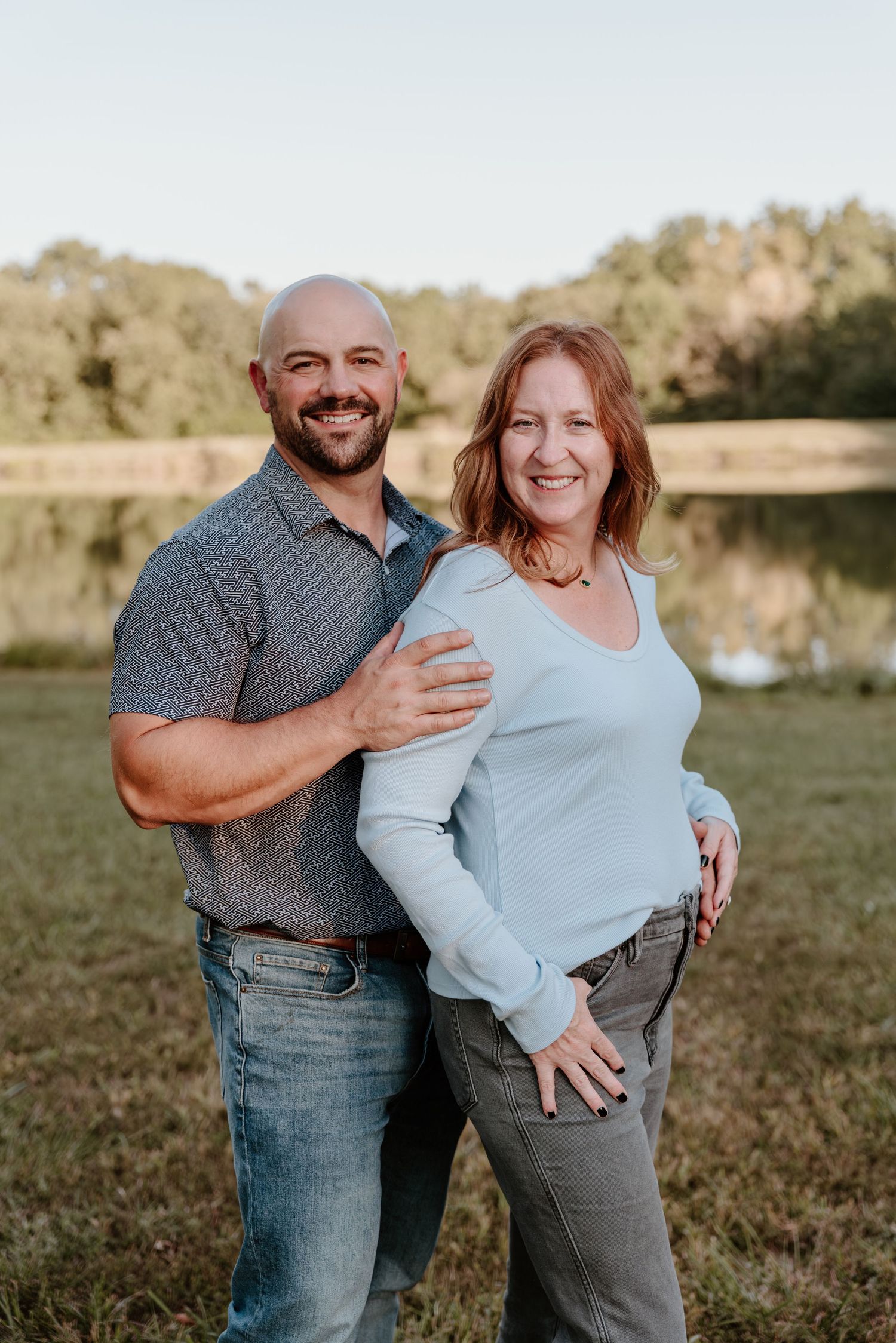 A couple poses together outdoors by a lake at sunset in casual attire on grassy terrain.