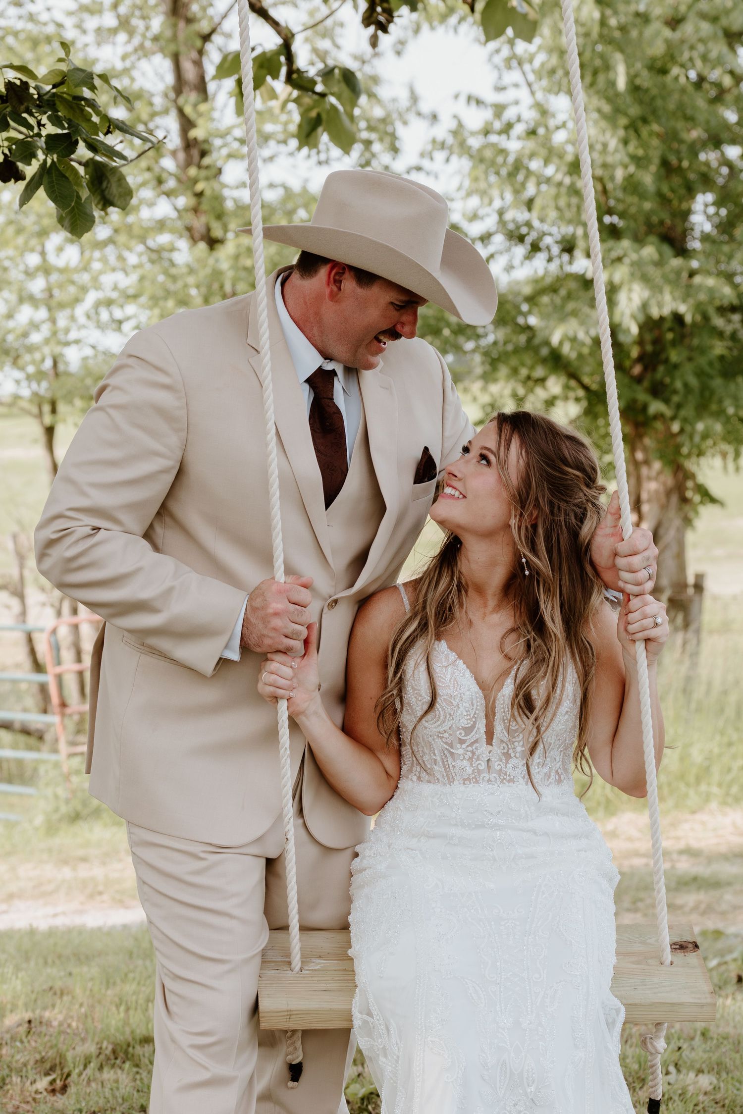 Romantic bride and groom in tan suit and cowboy hat sharing a tender moment on a wooden swing outdoors.