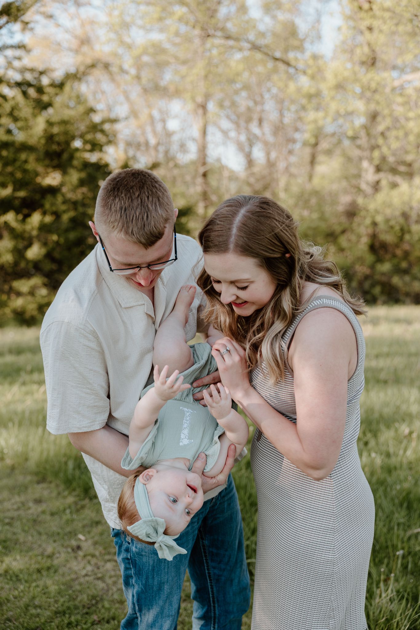 A young family smiles and plays with their baby outdoors in a green park during a warm family portrait session.
