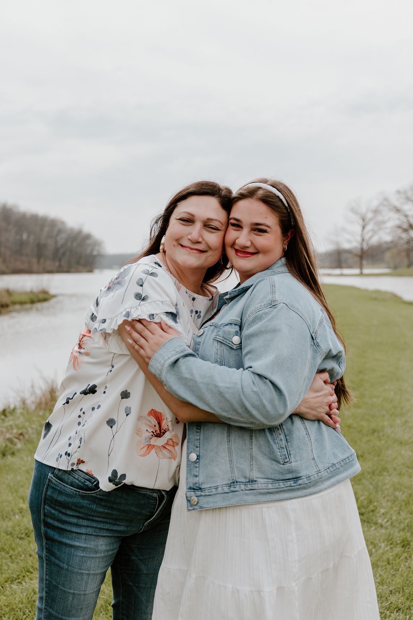 Two women hugging and smiling outdoors near a river, one in a floral blouse and jeans, the other in a denim jacket and white skirt.