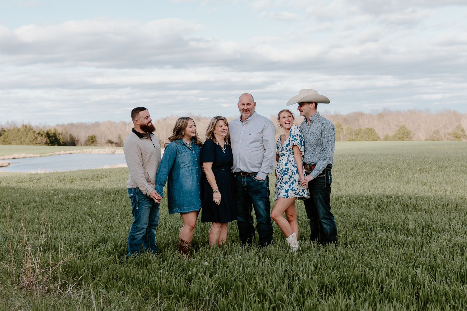 Family of six smiling together in a green field with mountains and cloudy sky in the background.