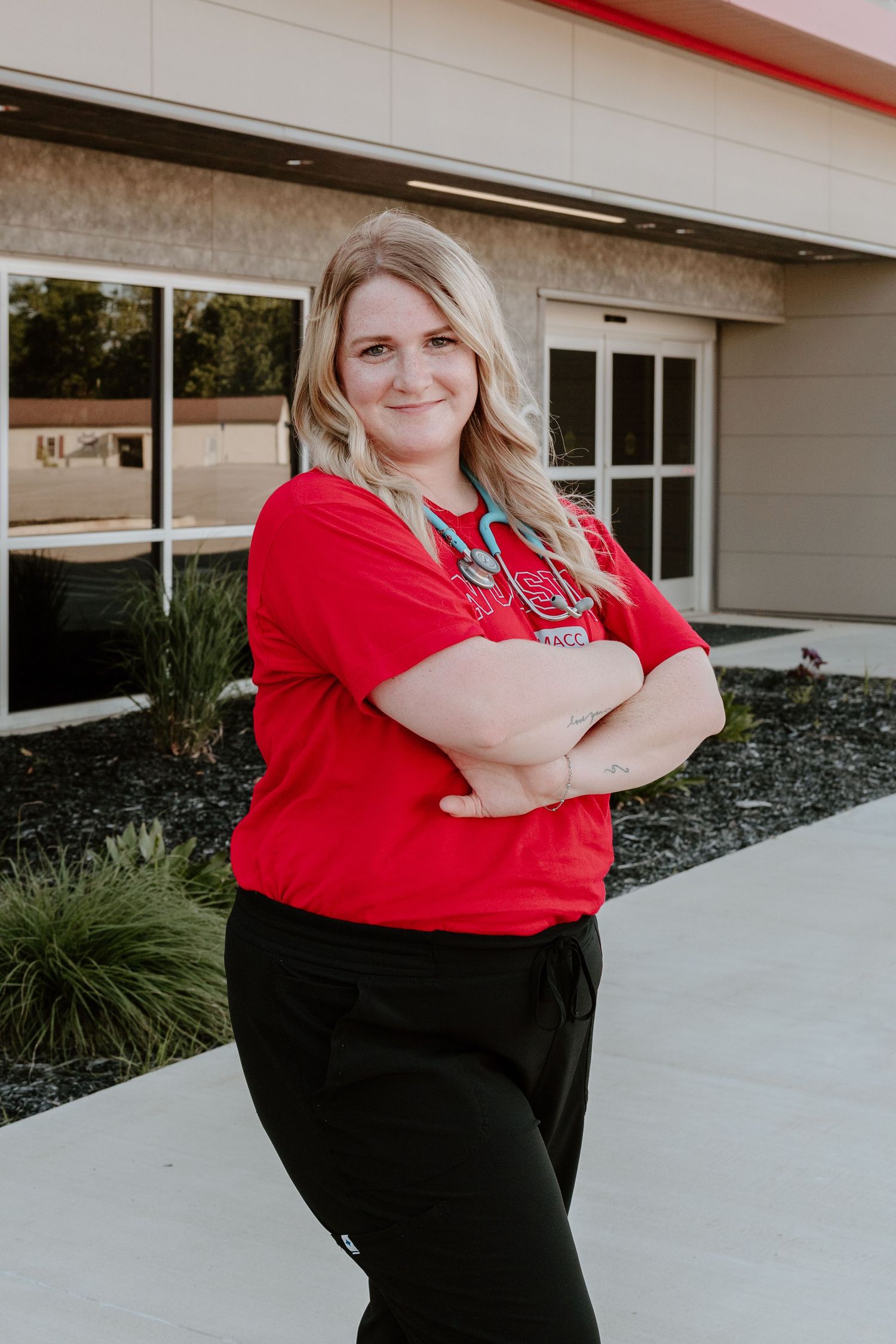 Healthcare worker in red scrubs and black pants stands confidently with arms crossed outside a medical building.